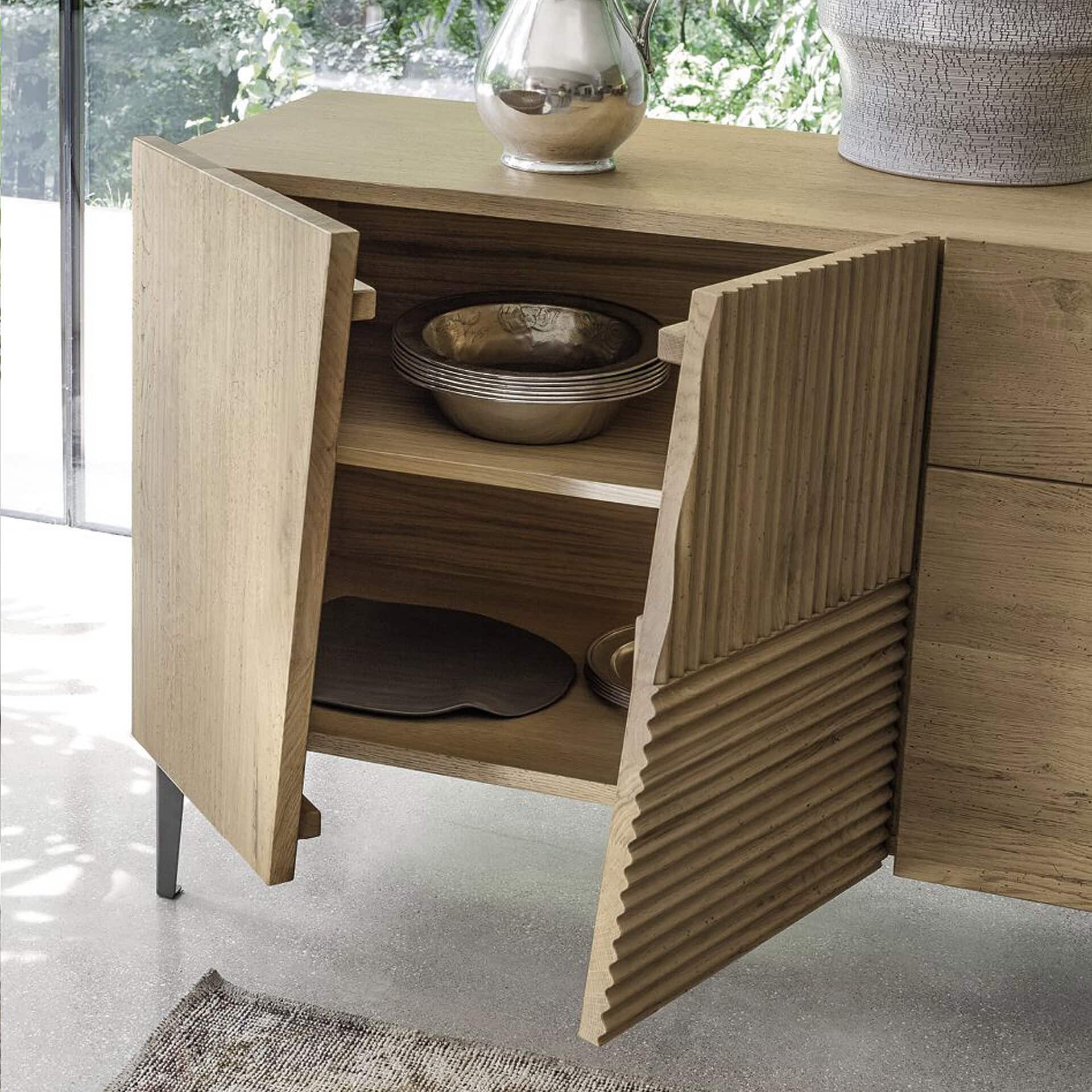 A modern light wood cabinet with two open doors. The right door features a unique fluted texture. Inside are decorative bowls and platters. A silver pitcher and textured vase sit on top. A window with green foliage is in the background.