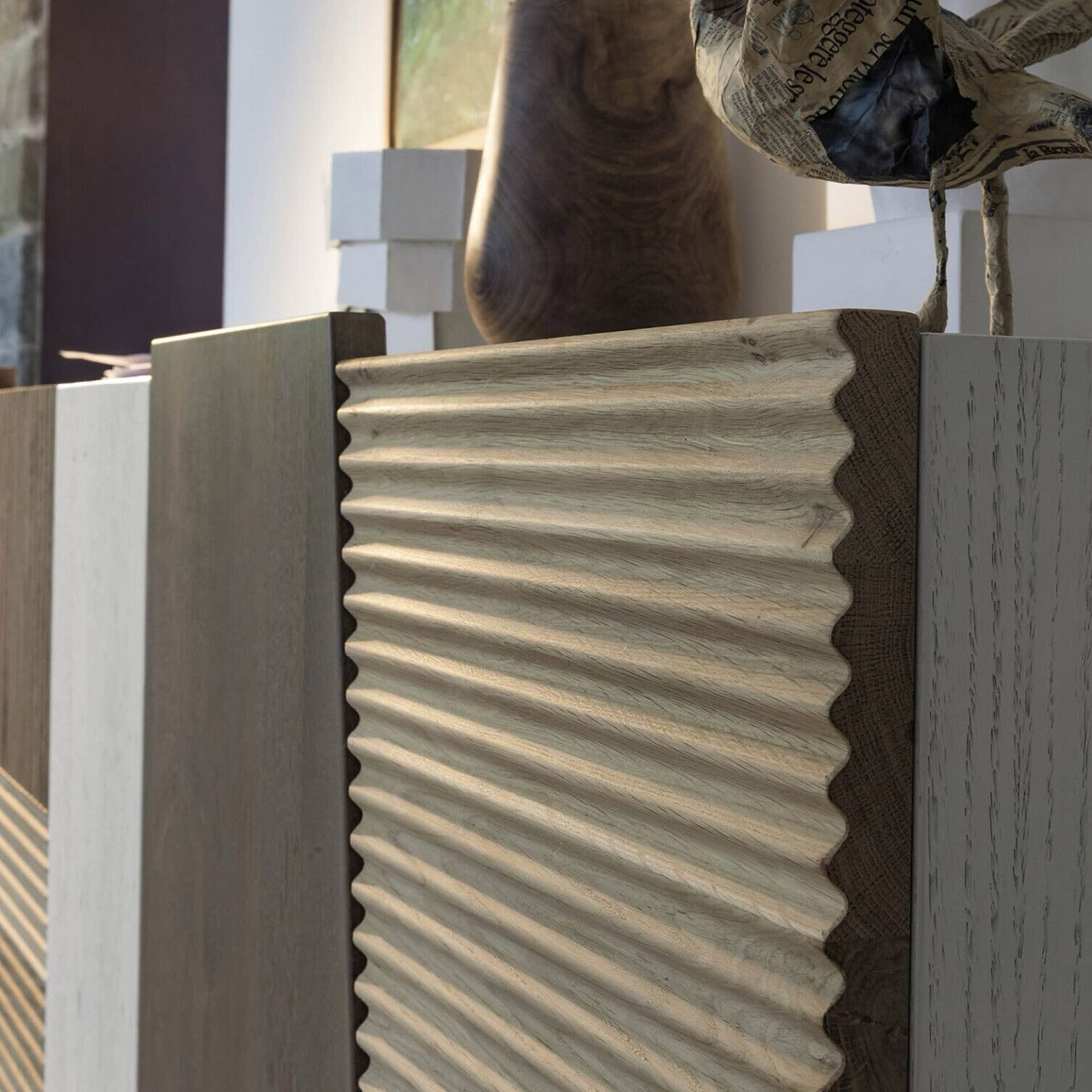 Close-up of a modern cabinet featuring a prominent light wood panel with a wavy, corrugated texture. It is flanked by other vertical wood panels in varying shades of brown and grey, and topped with a dark wooden vase and a bird sculpture made from newspaper.