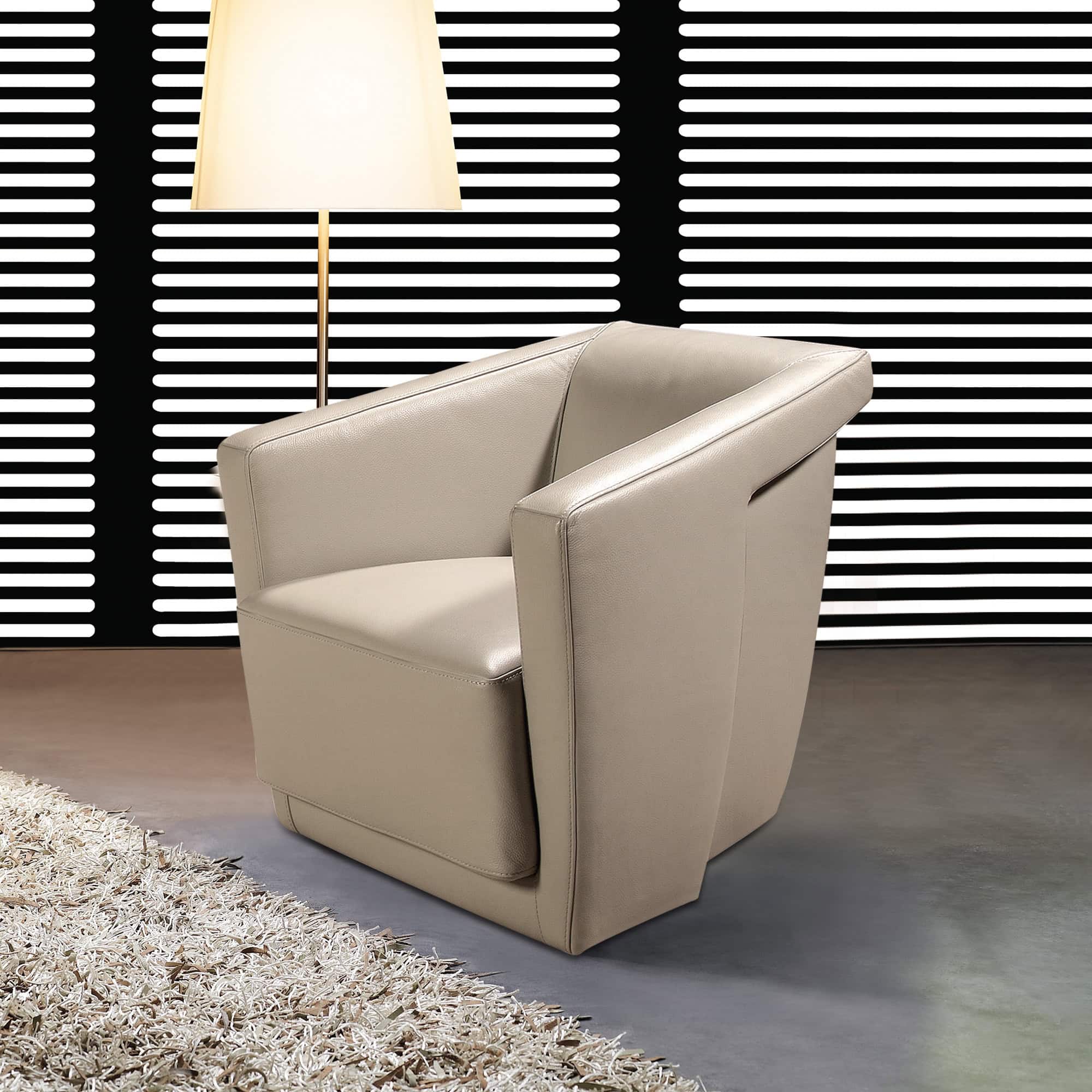 A light beige leather armchair with a modern, angular design. It sits on a dark floor next to a shaggy cream rug, with a tall floor lamp and a black and white striped wall in the background.