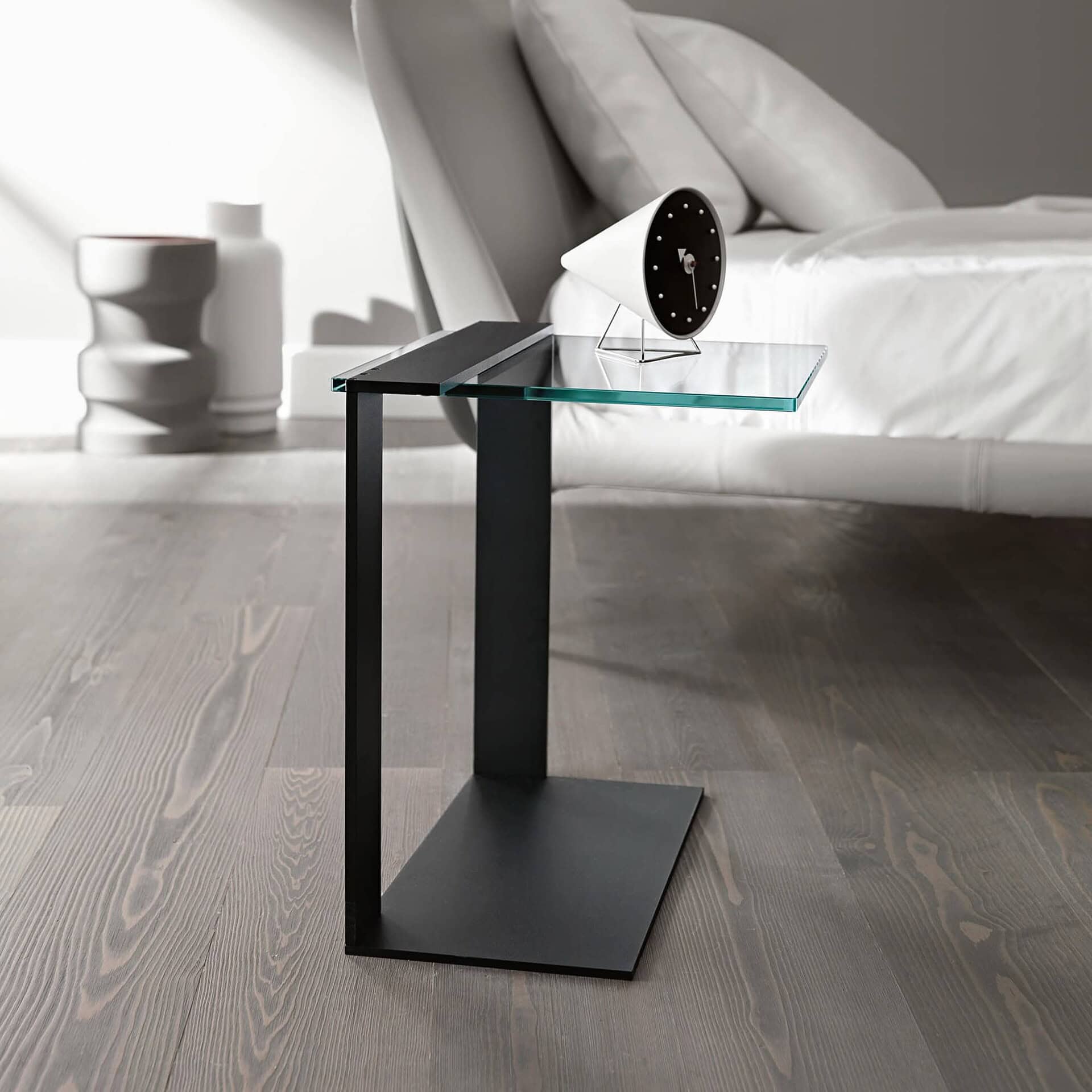 Black metal Joliet Side Table with angled glass top, placed beside a modern bed on a wooden floor.