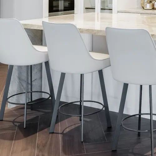 Three white bar stools with dark metal legs in front of a light marble kitchen island on a dark brown patterned floor.