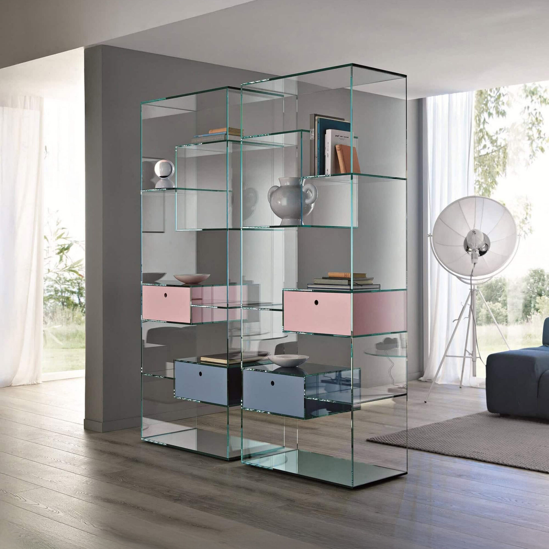 A modern, transparent glass bookshelf with pink and blue drawers, serving as a room divider in a bright, minimalist living space with wooden floors and large windows.