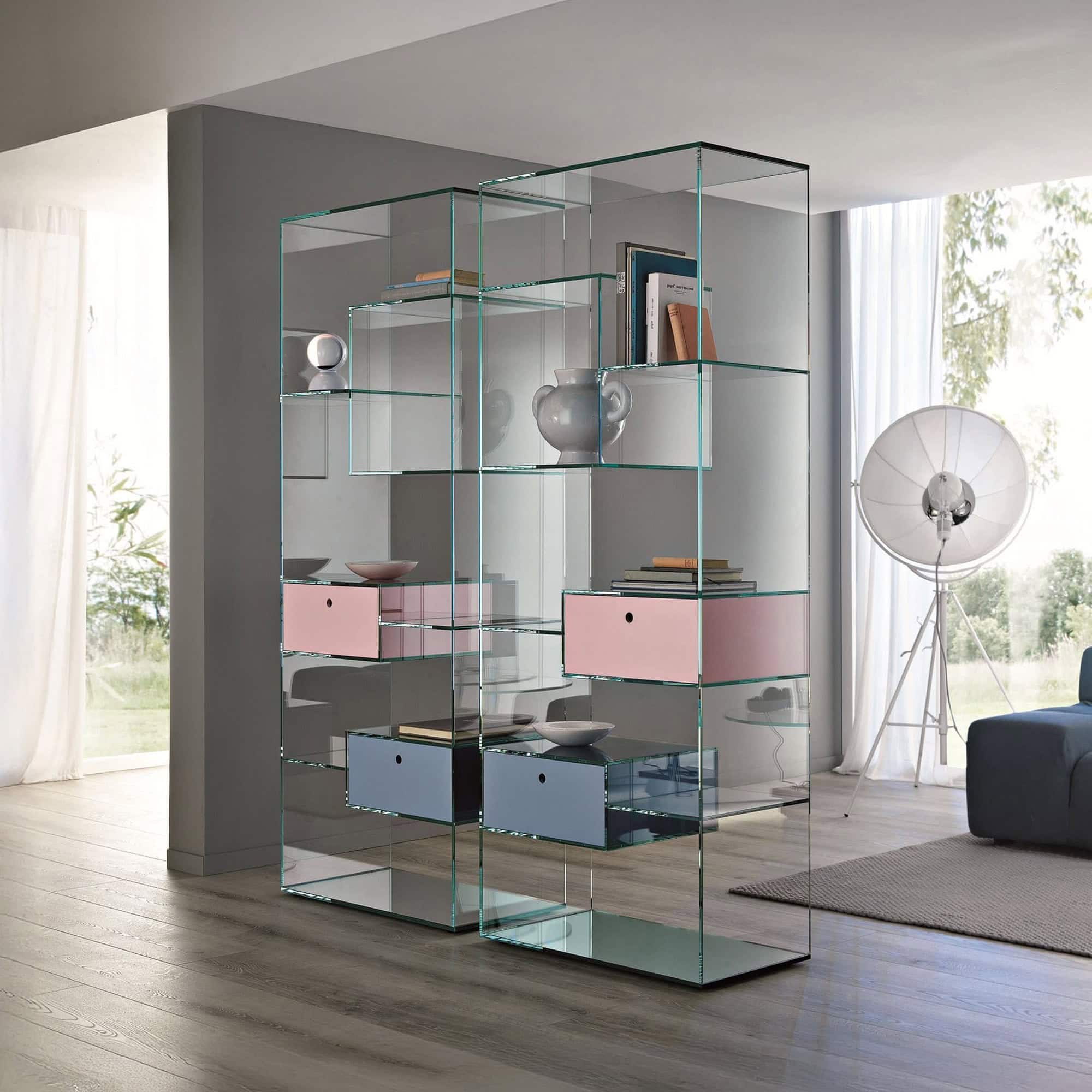 A modern, transparent glass bookshelf with pink and blue drawers, serving as a room divider in a bright, minimalist living space with wooden floors and large windows.