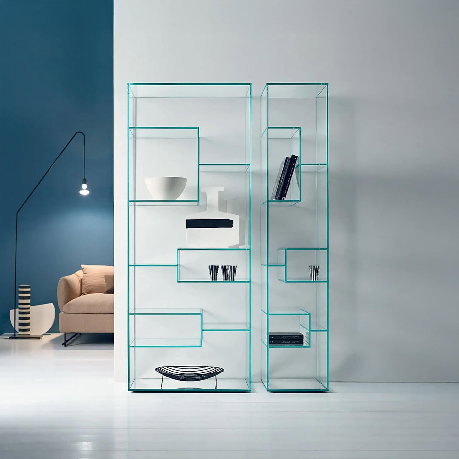 Two modern glass shelving units with asymmetrical shelves, displaying minimalist decor against a blue and light gray wall. A black floor lamp and a beige sofa are visible on the left.