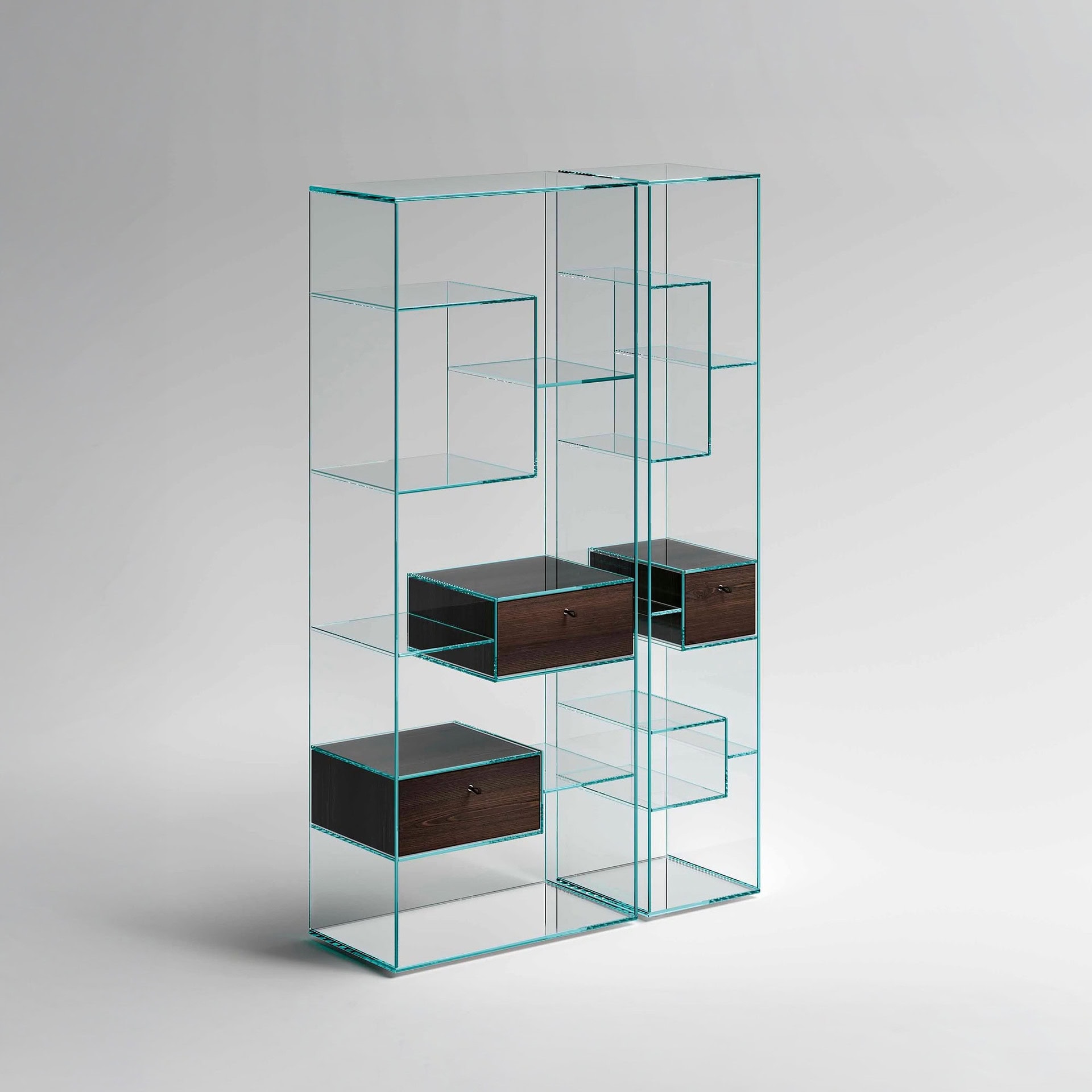 A modern glass bookshelf with a greenish tint on its edges, featuring multiple shelves and three dark wooden drawers with small handles, all against a light gray background.
