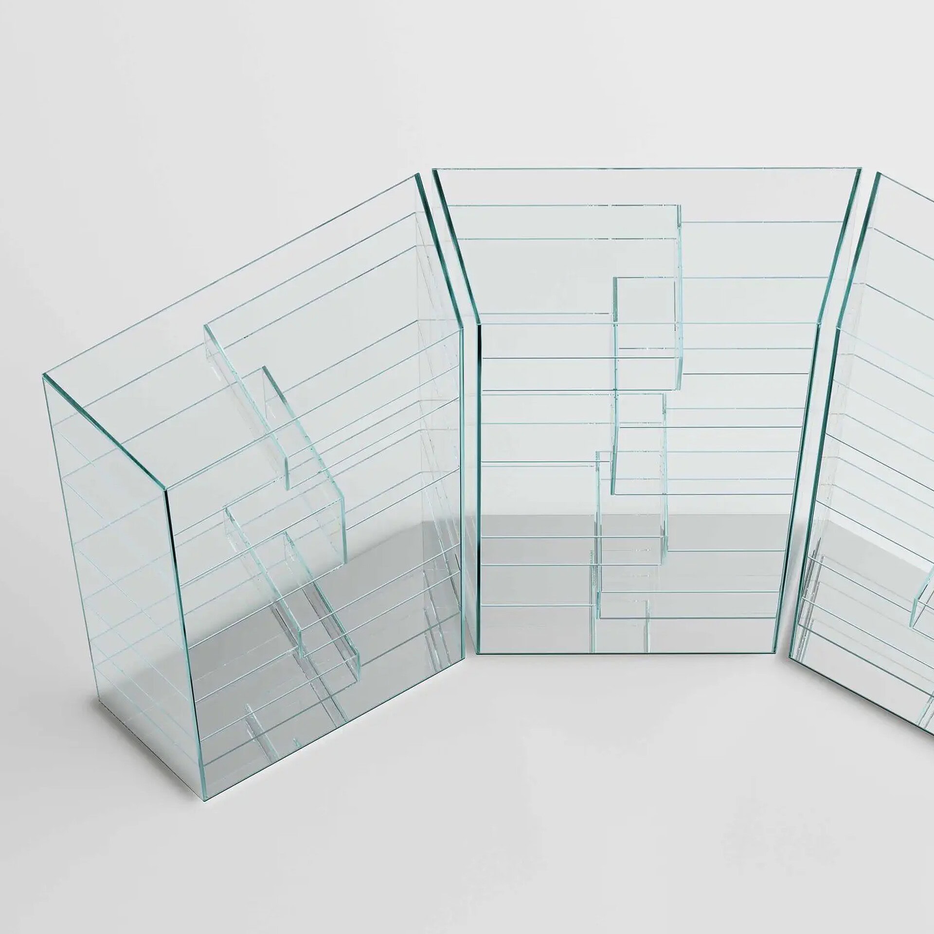 Three interconnected, modular clear glass display units with multiple shelves and compartments, arranged in a zig-zag pattern on a light gray surface.