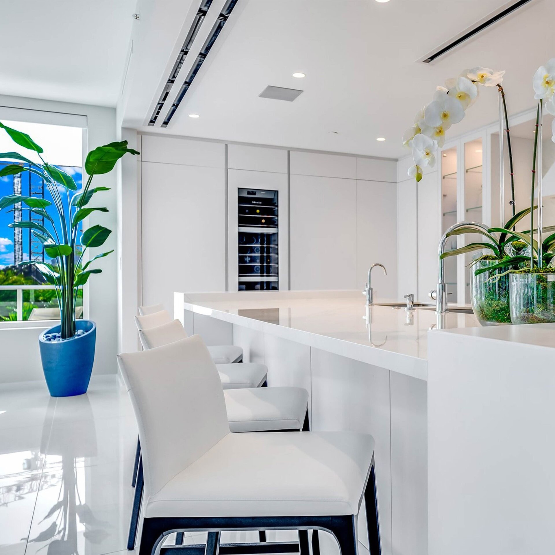 Modern white kitchen with sleek cabinetry and white island counter, featuring elegant barstools and lush green plants.