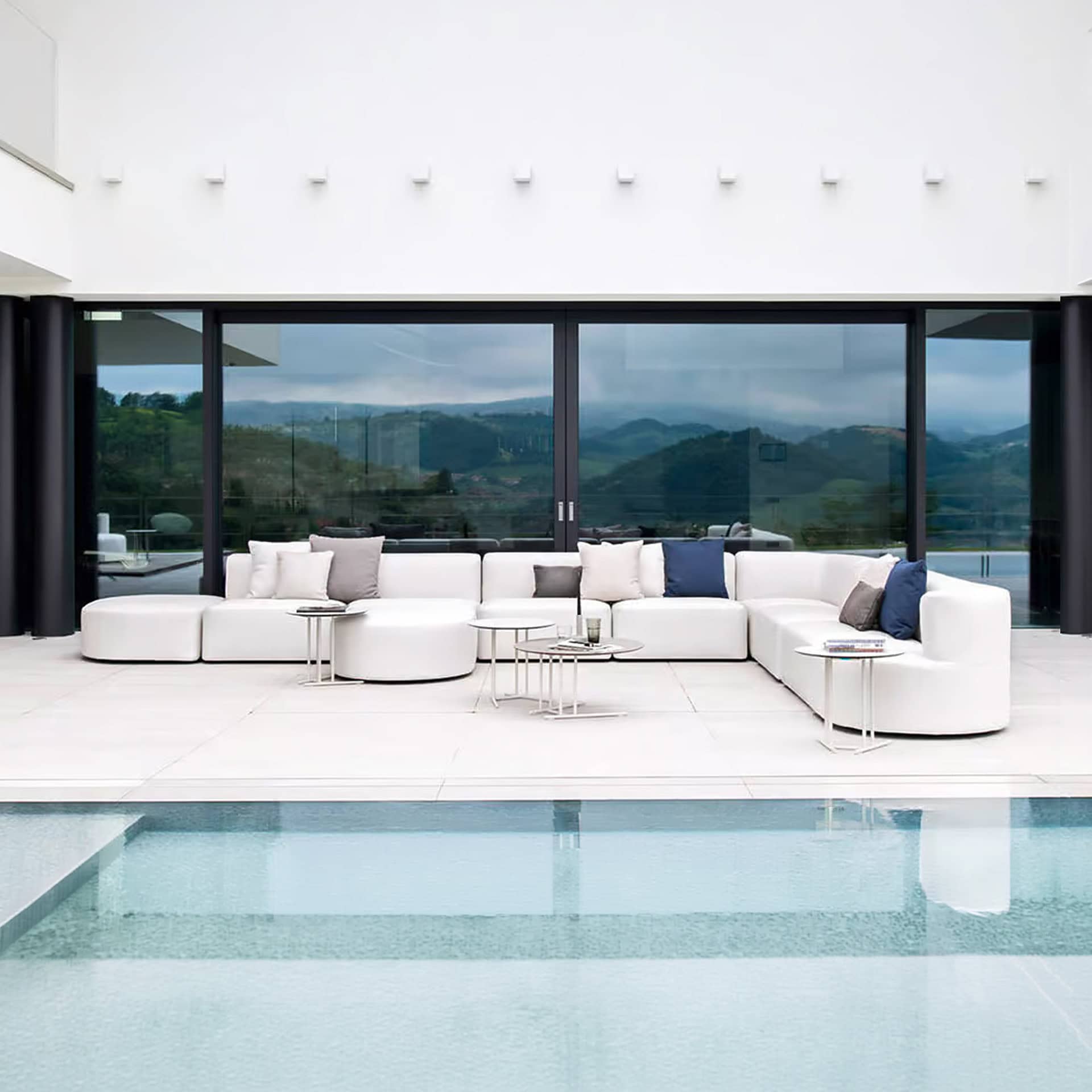 A modern outdoor patio with a large white modular sofa, small side tables, and a clear blue swimming pool. In the background, large glass doors overlook rolling green mountains under a cloudy sky.
