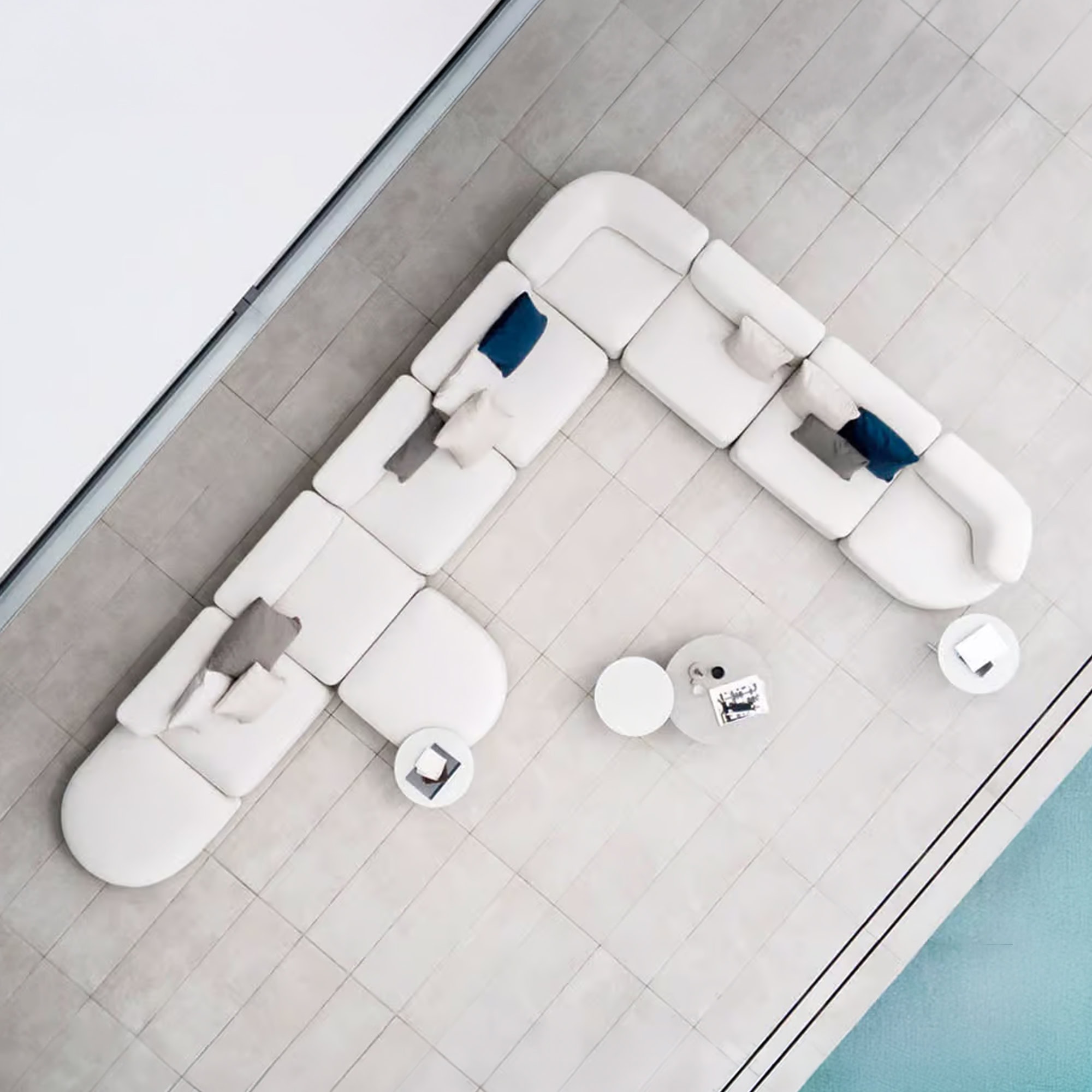 Overhead view of a large, L-shaped modular white sofa with blue and grey throw pillows on a light grey tiled patio, next to a turquoise swimming pool. Three small round white side tables are also visible.