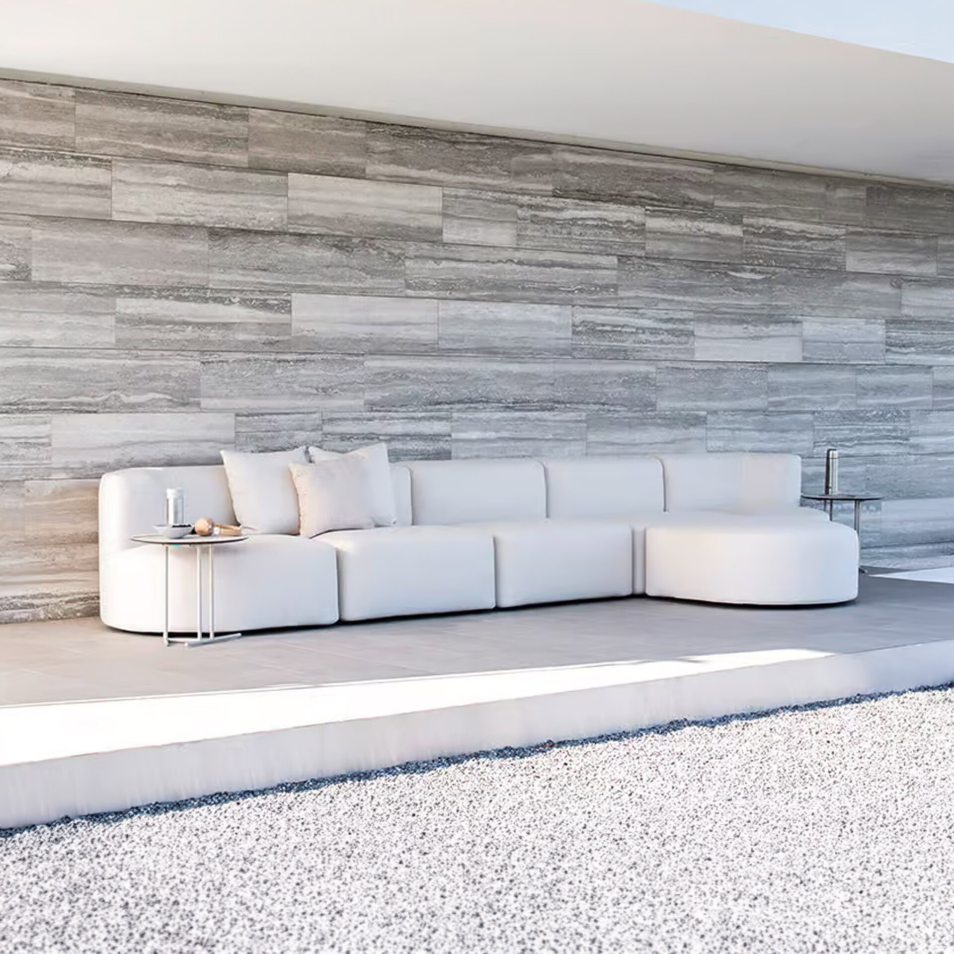 A luxurious L-shaped white modular sofa with throw pillows in a modern outdoor setting. The sofa is placed against a textured grey stone-tiled wall, with light grey tiled flooring and a foreground of white gravel. Two small glass-top side tables with drinks and snacks flank the sofa.