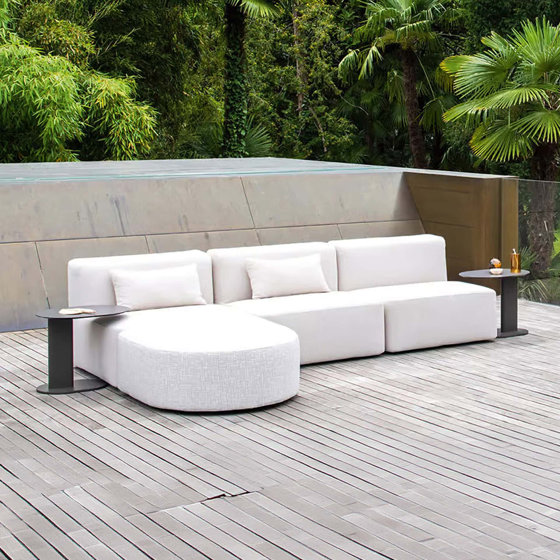 An L-shaped white modular outdoor sofa with side tables on a wooden deck, backed by a modern wall and lush green tropical foliage.