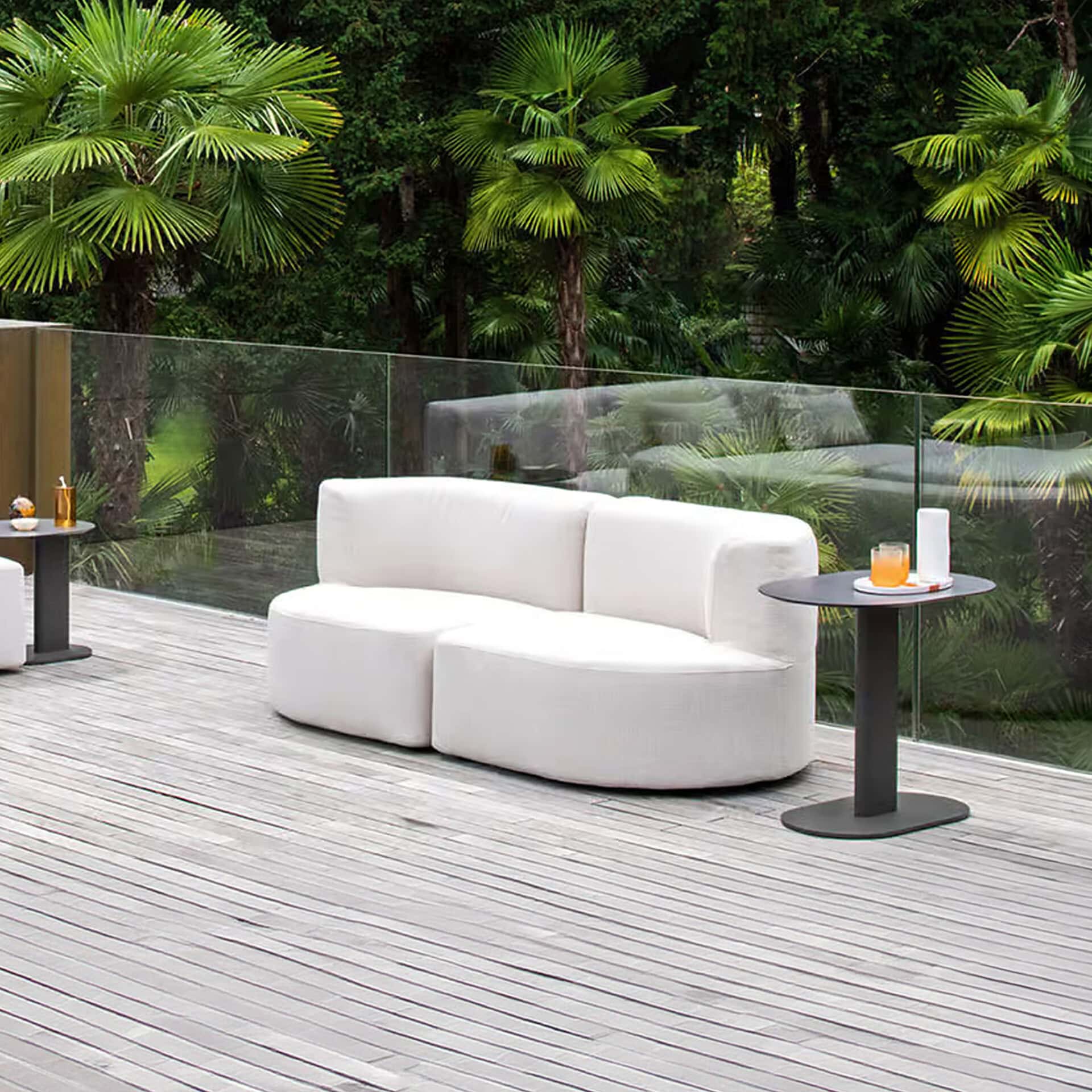 A modern white outdoor sofa on a light wooden deck, with dark side tables, overlooking a lush garden of palm trees through a clear glass railing.
