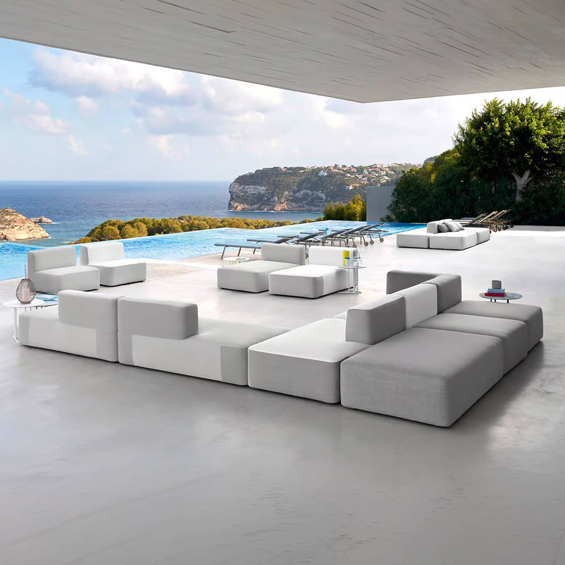 A modern outdoor patio with modular white and grey seating, an infinity pool, and a panoramic ocean view with cliffs and a distant town under a blue sky.