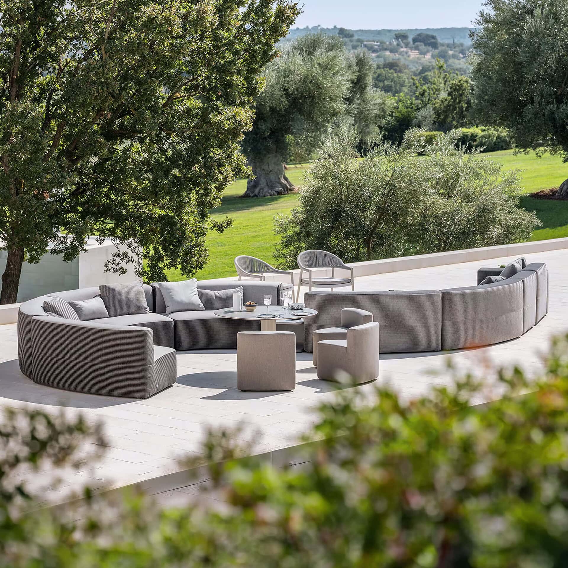 A luxurious outdoor patio featuring a large curved modular sofa, a round coffee table, and armchairs on light-colored tiles. The setting is surrounded by lush green trees, including ancient olive trees, and rolling hills under a clear blue sky.