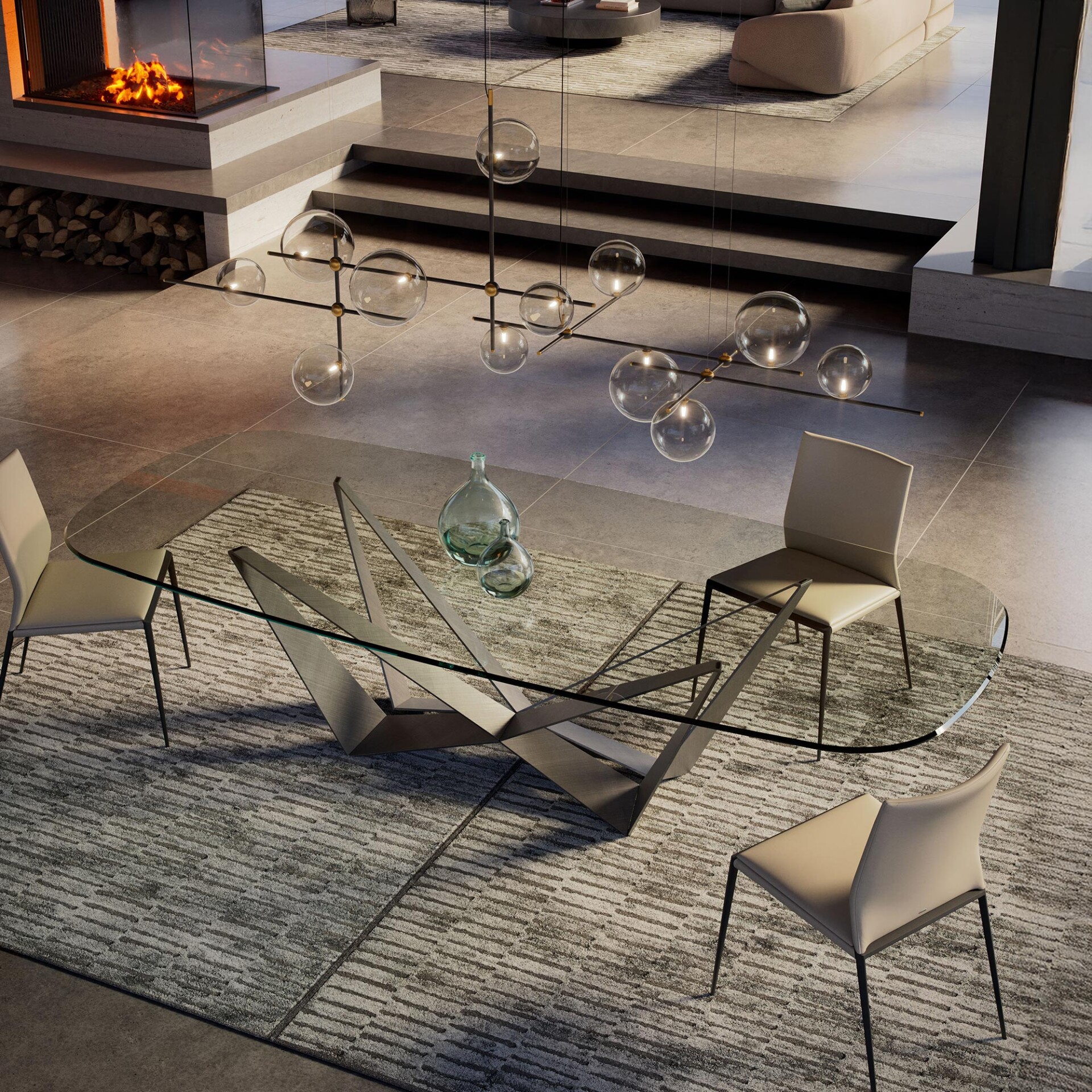 A luxurious modern open-plan living and dining area. In the foreground, a glass dining table with a geometric metal base and four beige chairs sits on a patterned rug. Above, a linear chandelier with glass globes hangs. In the background, steps lead to a raised living area with a modern fireplace and a light-colored sofa.