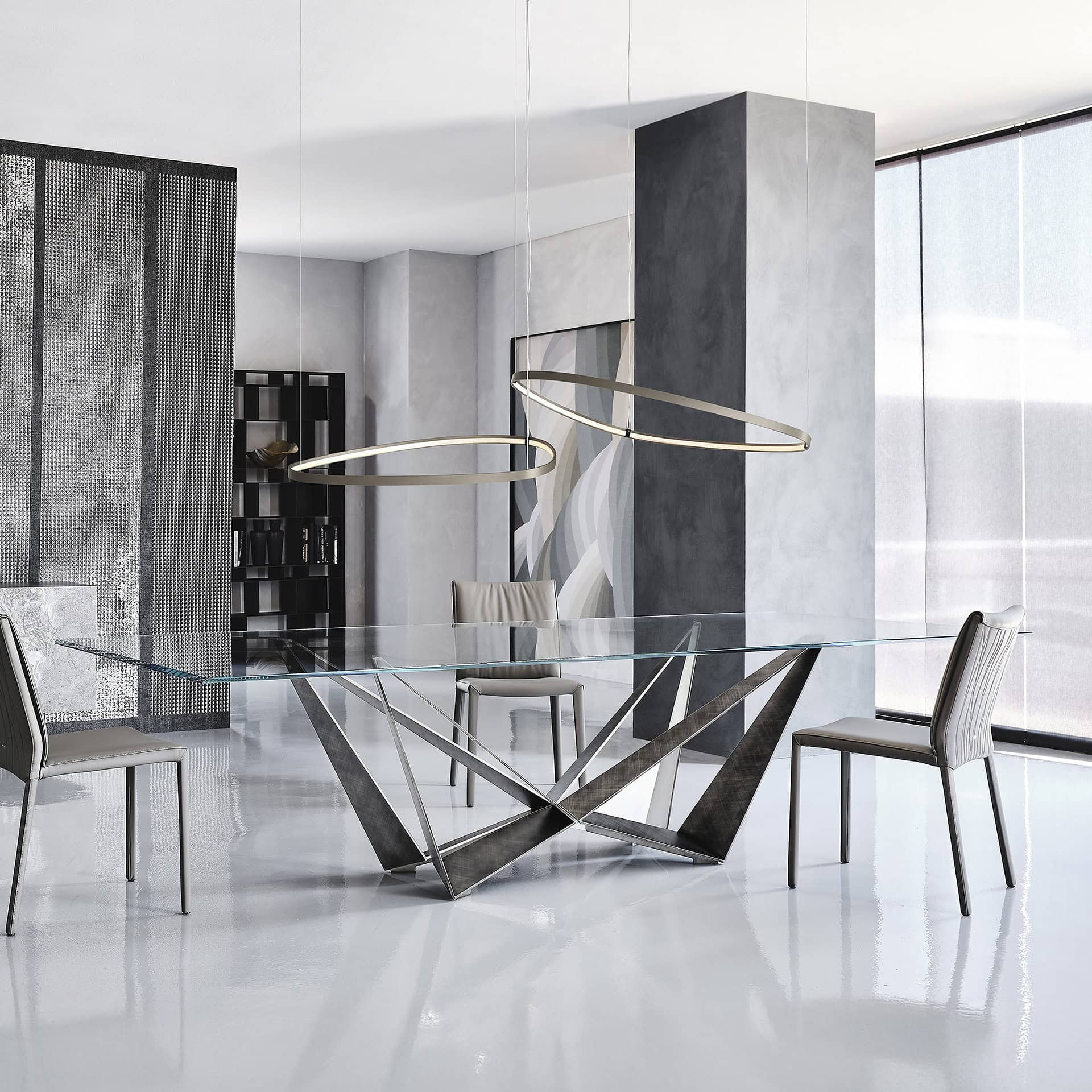 A modern dining room with a rectangular glass table featuring a sculptural dark metal base, surrounded by gray upholstered chairs. Two oval pendant lights hang above the table. The room has gray walls, a textured screen, and a large window with sheer blinds.