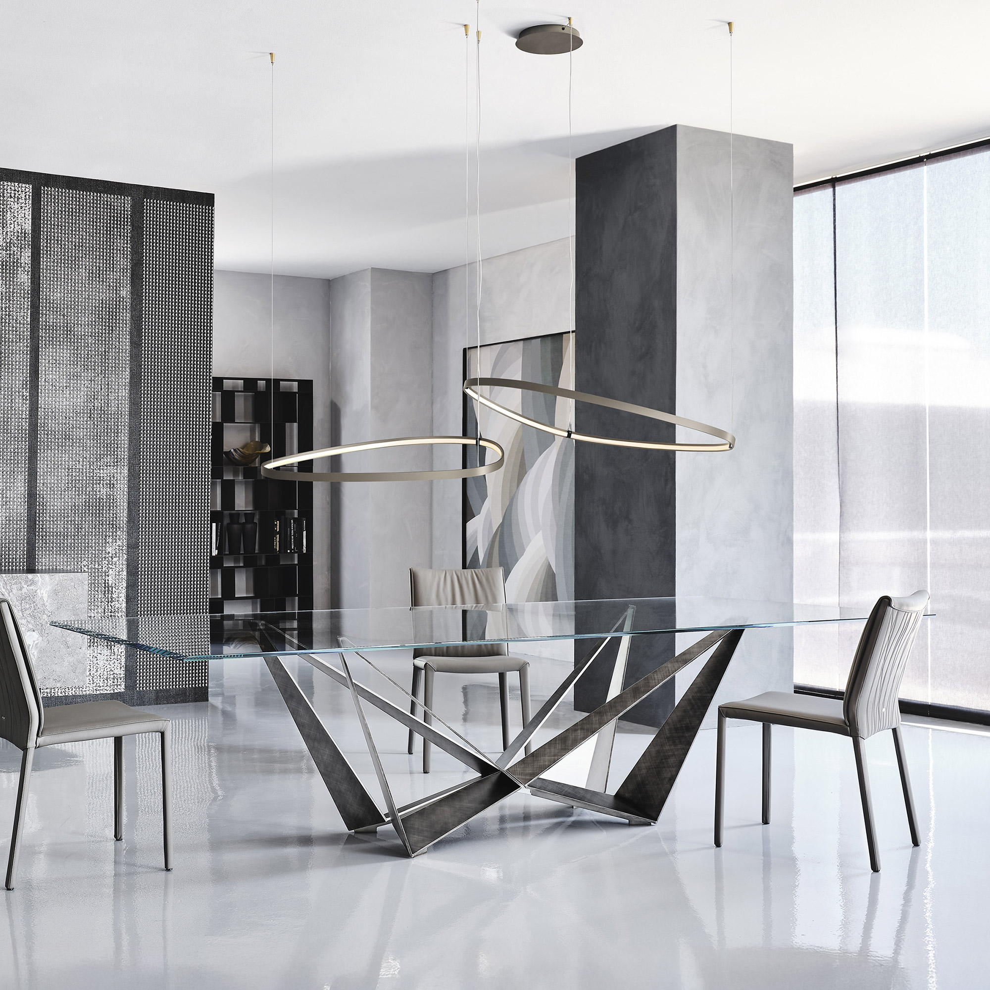 A modern dining room with a rectangular glass table featuring a sculptural dark metal base, surrounded by gray upholstered chairs. Two oval pendant lights hang above the table. The room has gray walls, a textured screen, and a large window with sheer blinds.