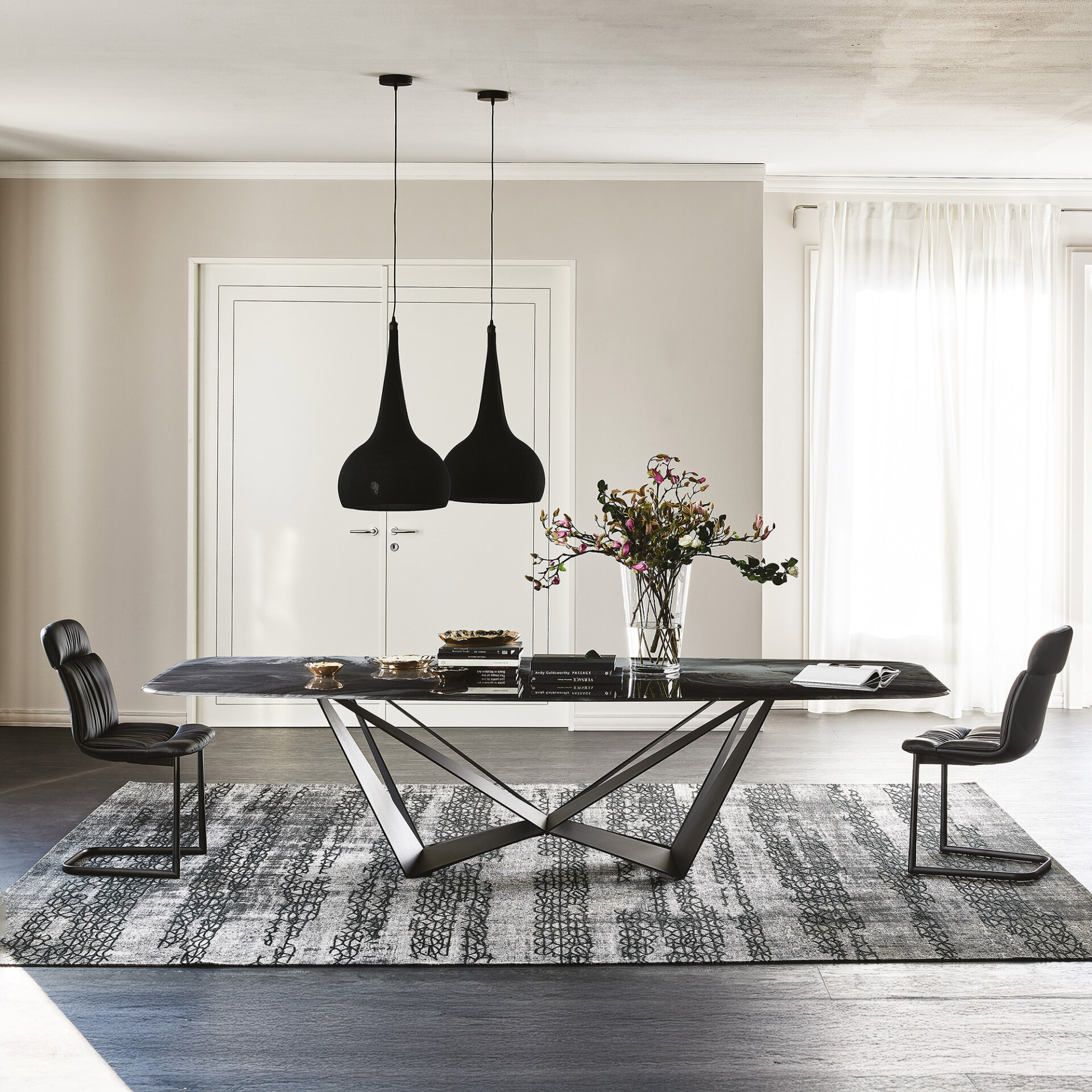 A modern dining room with a dark rectangular table, two black leather chairs, two black pendant lights, and a grey patterned rug. A vase of flowers and books decorate the table. White doors and sheer curtains are in the background.