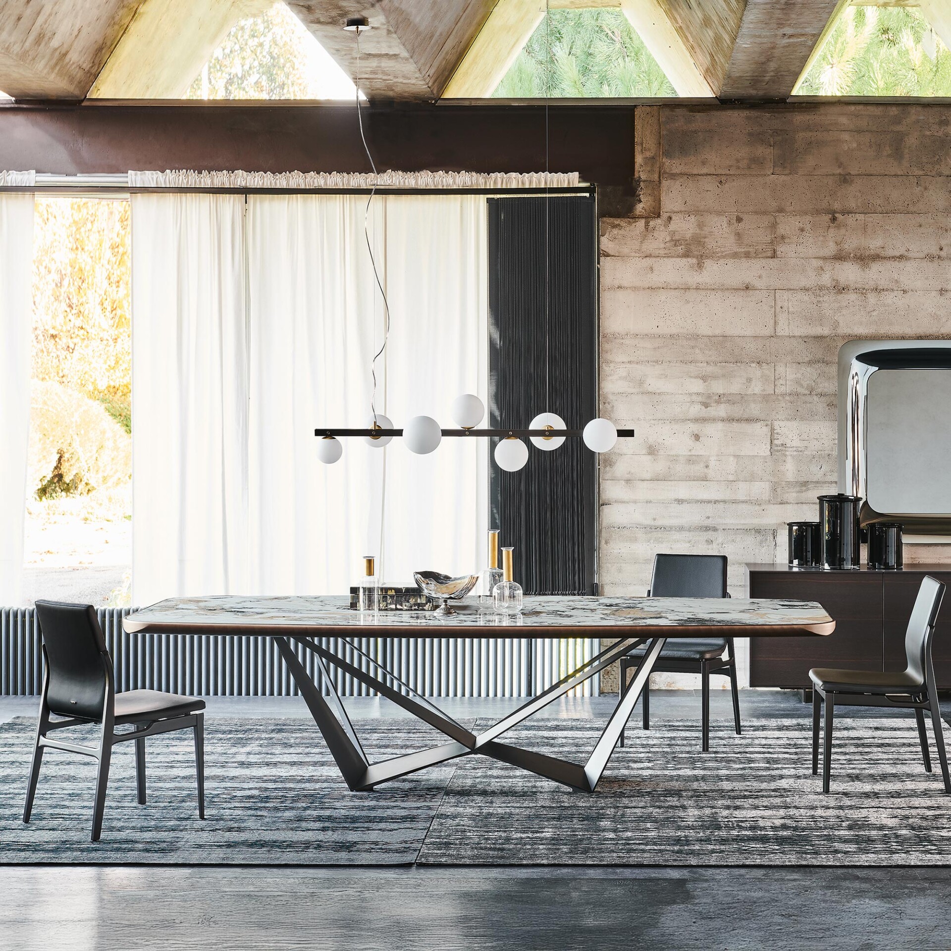 A modern dining room with a large marble-top table, dark chairs, and a linear globe chandelier. Exposed concrete walls and ceiling, sheer white curtains, and a patterned rug complete the industrial-chic aesthetic.