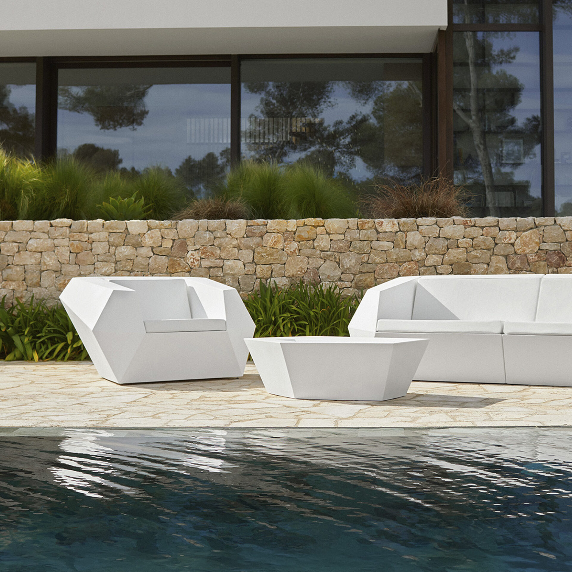 A white geometric outdoor armchair and a matching low table next to a swimming pool, with a stone wall and green plants in the background.