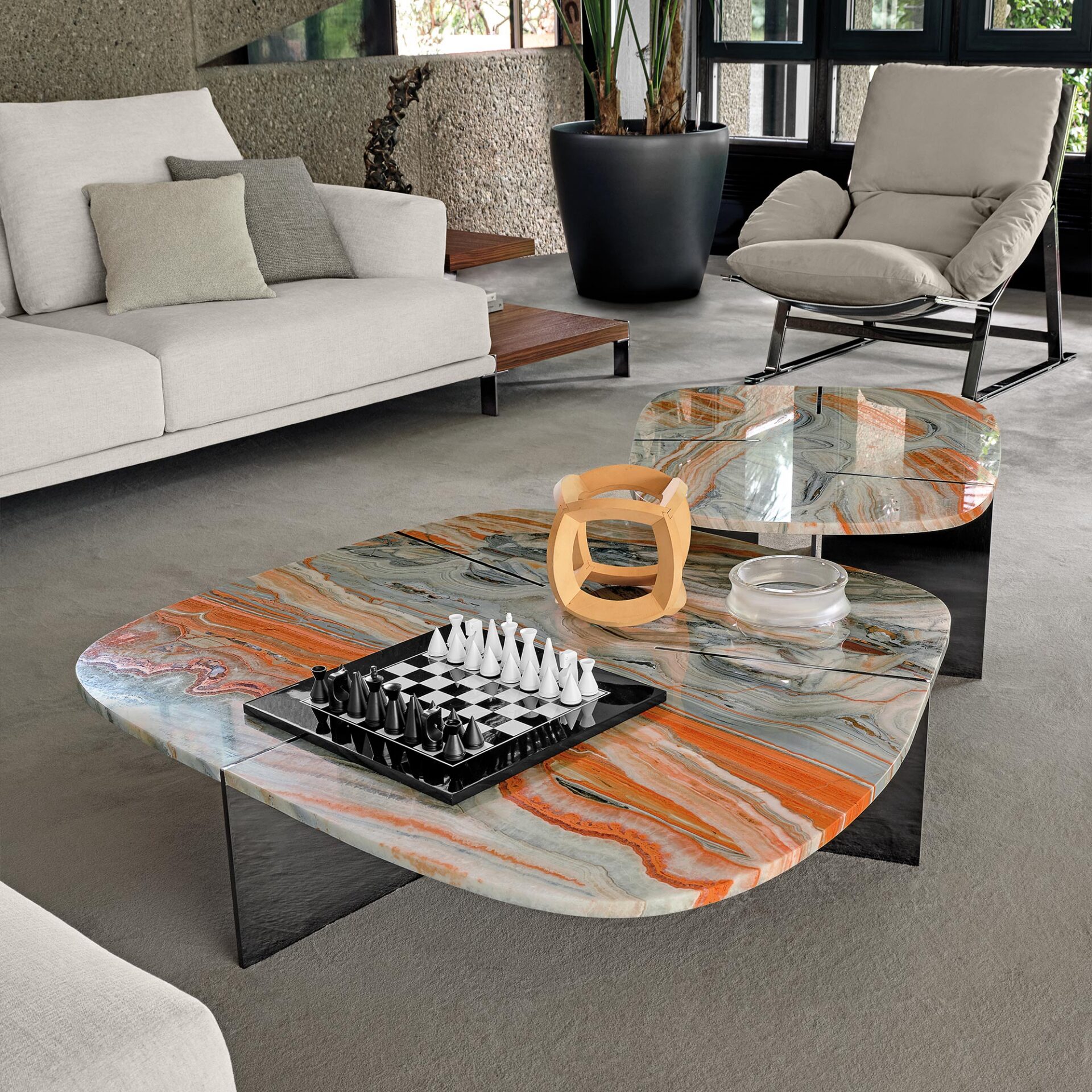 A modern living room with two vibrant orange and grey marble coffee tables, a light beige sofa, and an armchair. A chess set and a wooden sculpture are on the tables. Large windows and a concrete wall are in the background.