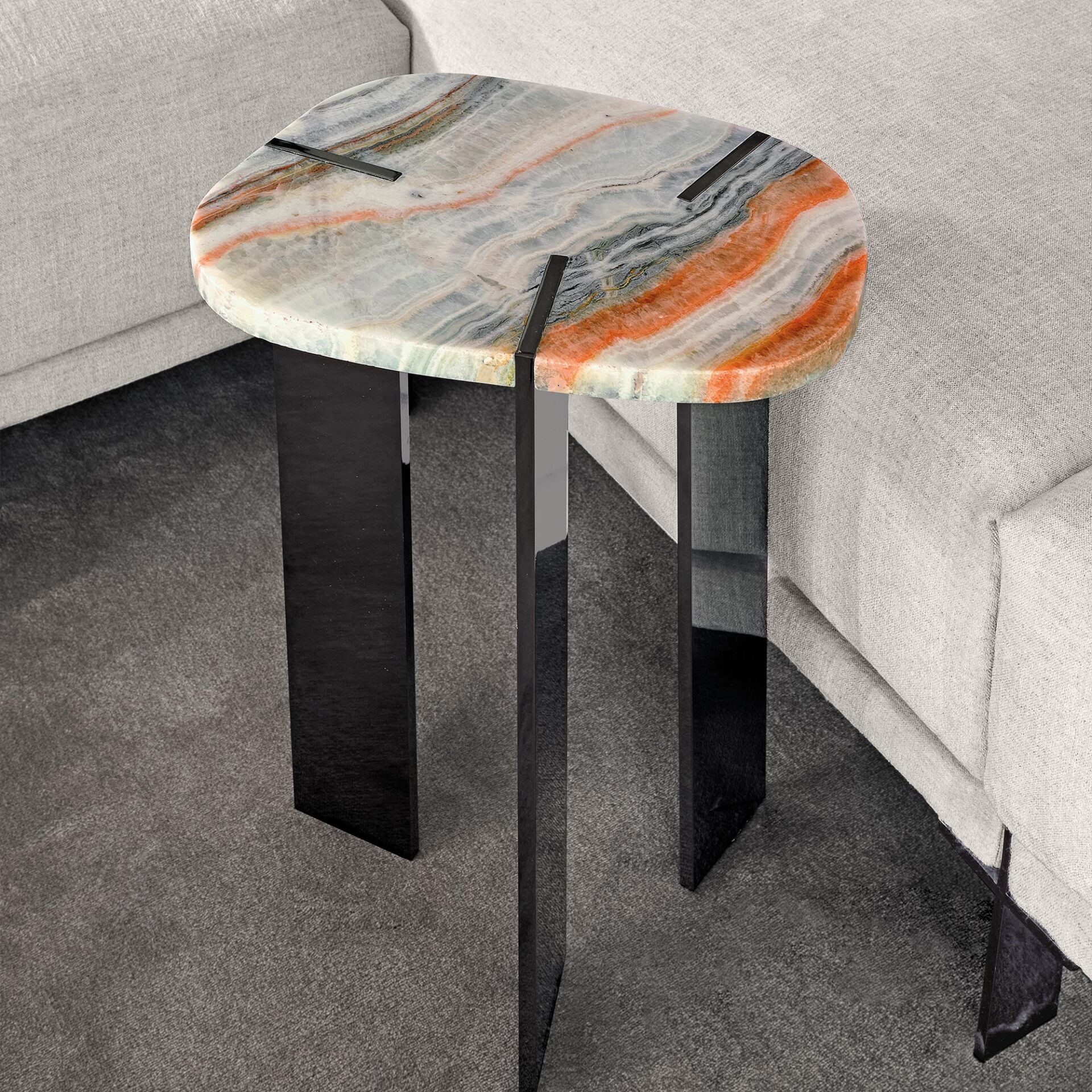 A modern side table with an irregular oval top made of colorful veined onyx in white, orange, and grey, supported by three sleek black rectangular legs. It stands next to a light grey sofa on a dark grey carpet.