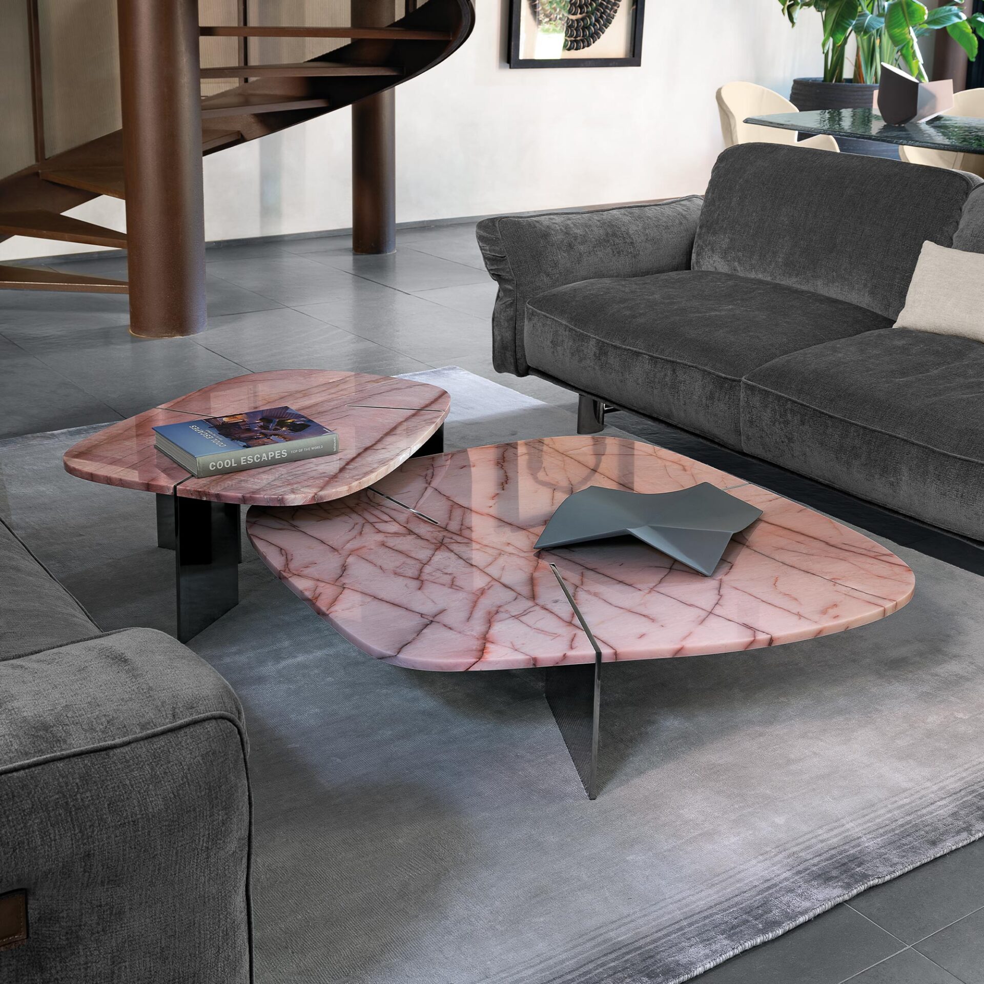 A modern living room with two irregularly shaped pink marble coffee tables on a grey rug, flanked by dark grey sofas. A dark metal spiral staircase is in the background.