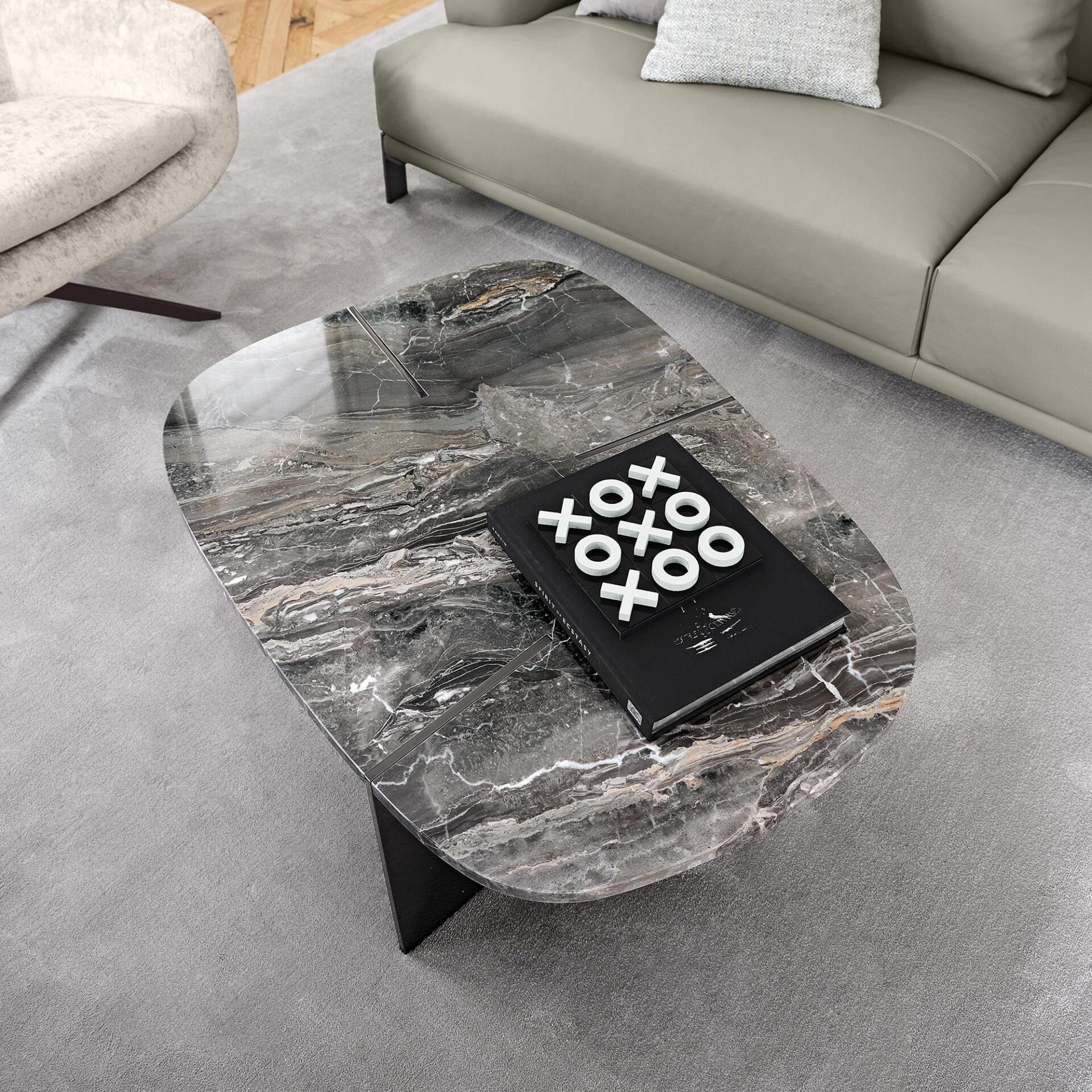 A modern living room featuring a dark marble coffee table with a tic-tac-toe game on top, a light grey rug, a taupe leather sofa, and a cream armchair.