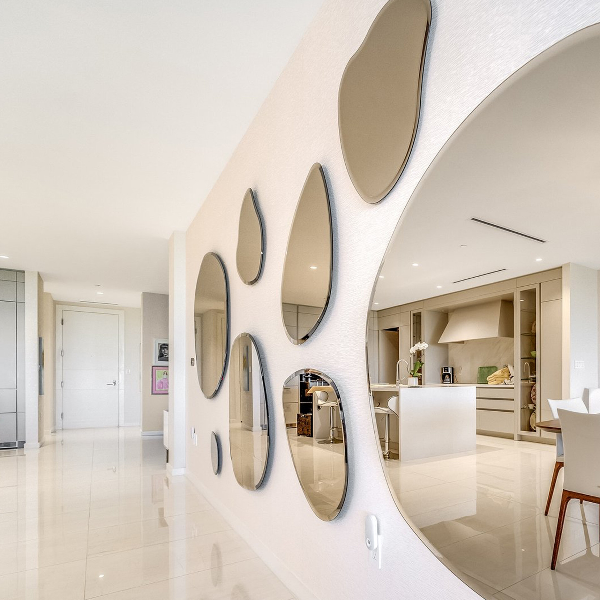 A modern interior with a hallway on the left and a textured wall adorned with multiple irregularly shaped, frameless mirrors on the right. The mirrors reflect a light-toned, contemporary kitchen and dining area.