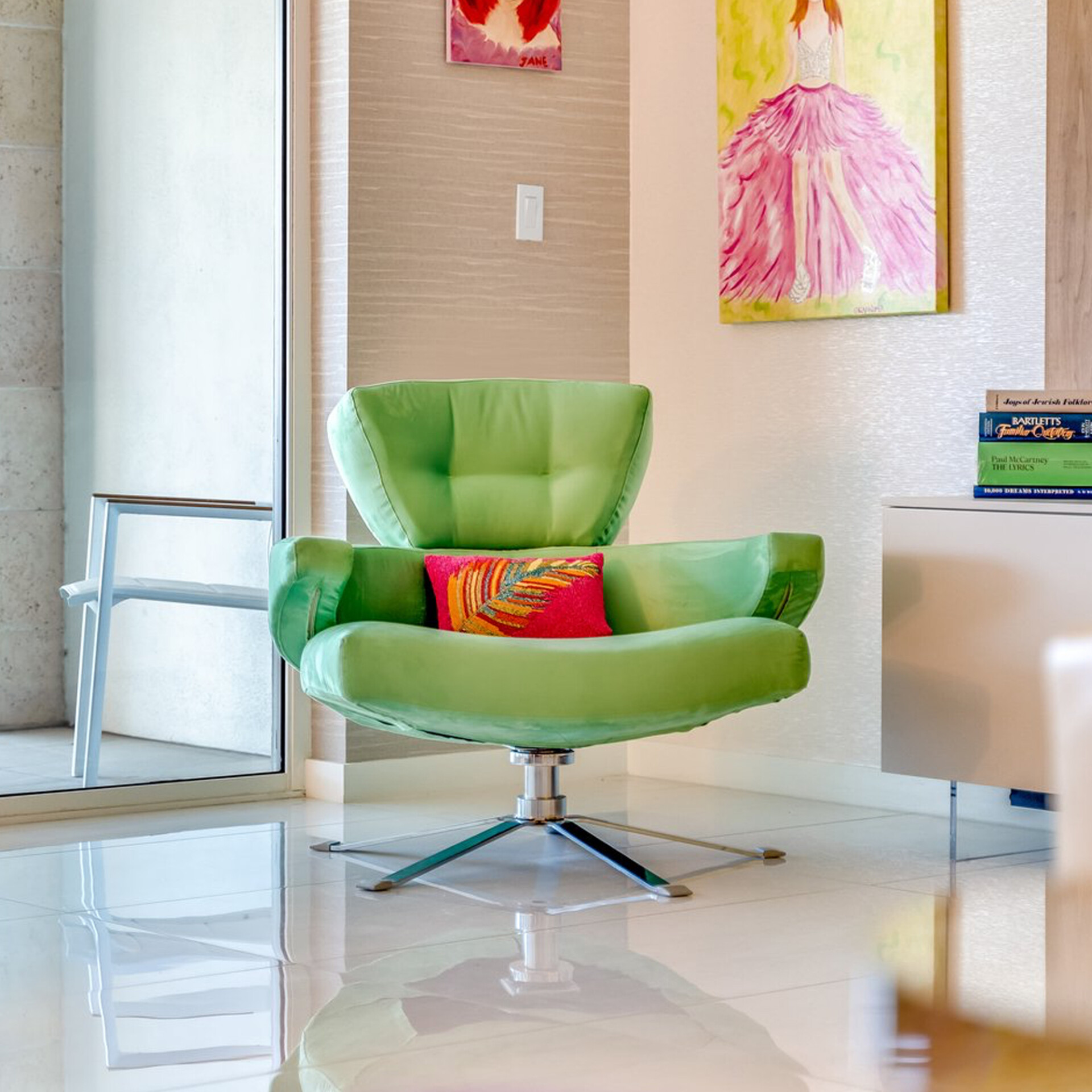 A bright lime green armchair with a colorful feather-patterned pillow sits on a white tiled floor in a modern room. The walls are adorned with eclectic artwork, and a stack of books rests on a white cabinet to the right.