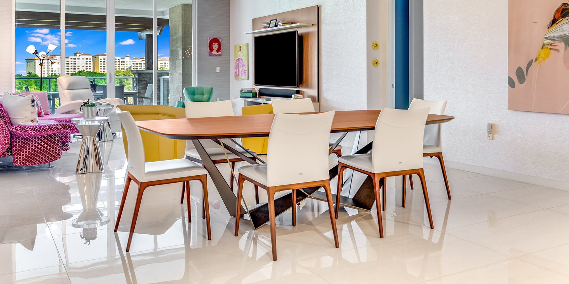 A modern, open-concept living and dining room with a vibrant color palette. A wooden dining table with cream chairs is in the foreground. A magenta sofa, yellow couch, and white recliner furnish the living area. A large TV is mounted on a wooden panel, and large windows offer a view of buildings, trees, and a blue sky.