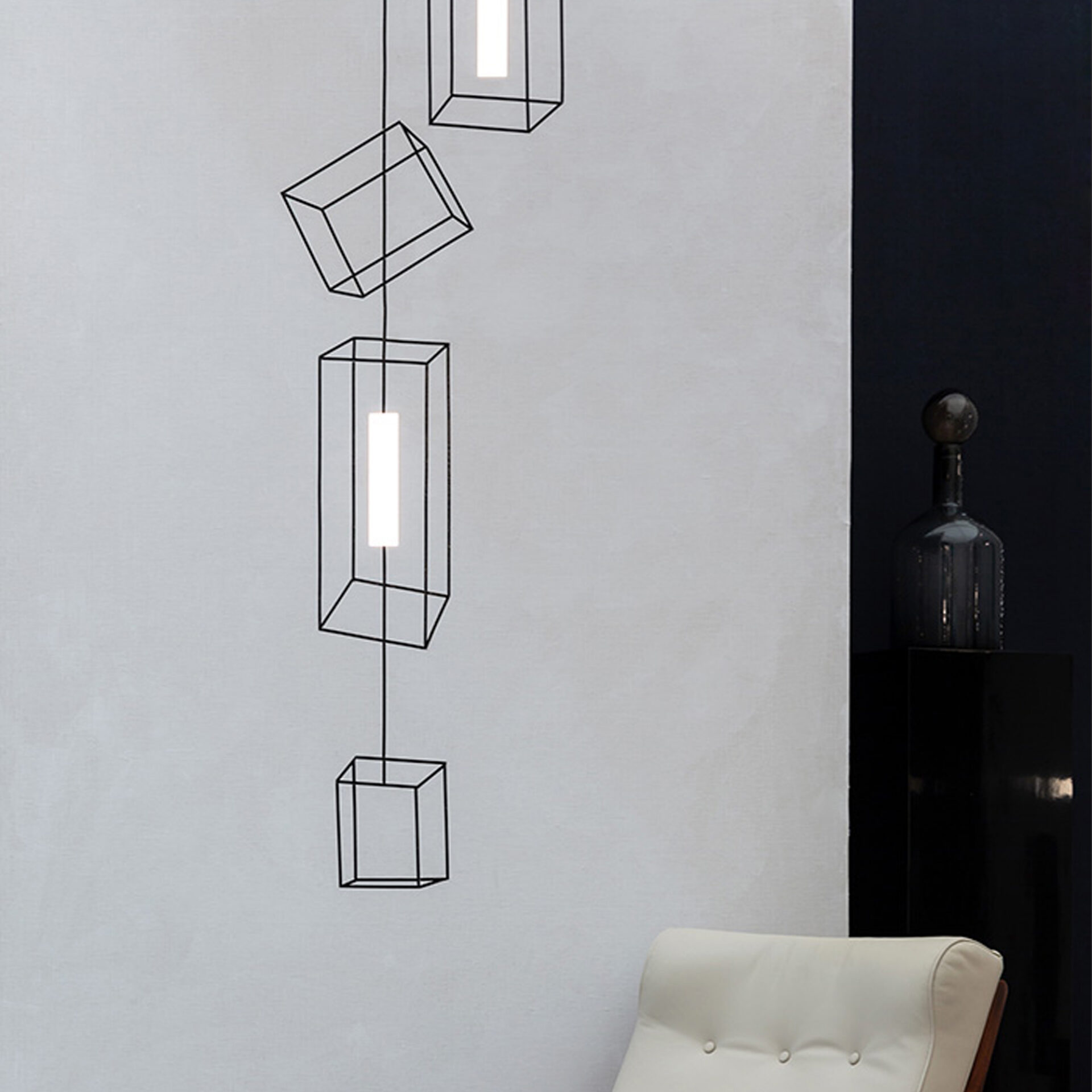 Modern interior featuring a vertical arrangement of black wireframe geometric pendant lights with glowing white elements against a light gray wall. A cream upholstered chair and dark decorative bottle are visible on the right.