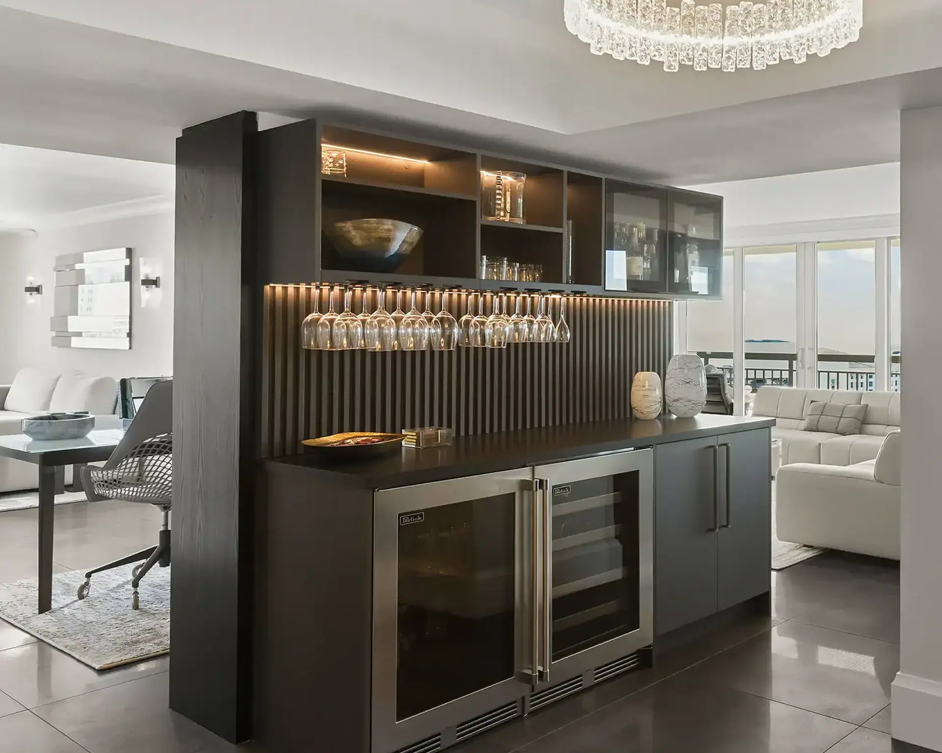 A modern built-in home bar with dark wood cabinetry, stainless steel wine coolers, and hanging wine glasses, separating a dining area from a living room with a white sofa and a view of an outdoor balcony.