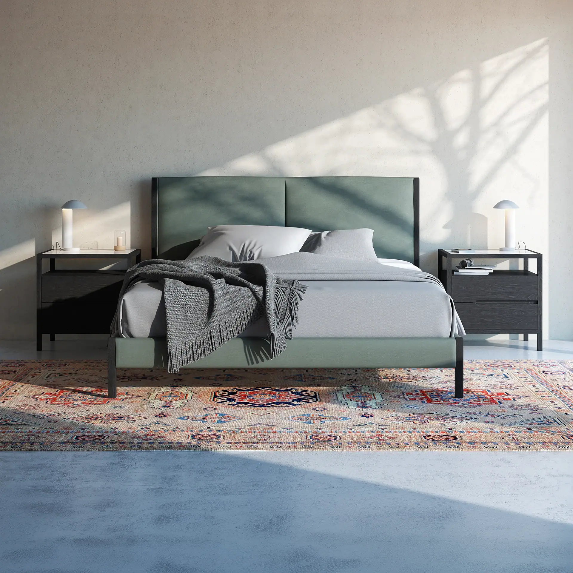 A modern bedroom with a sage green bed, gray bedding, dark gray nightstands with lamps, and a colorful oriental rug on a concrete floor, illuminated by natural light and shadows.
