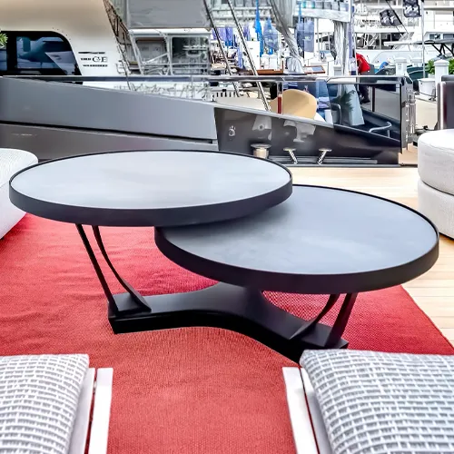 A modern two-tiered circular coffee table with dark gray tops and a black base, sitting on a red rug. A gray yacht is visible in the background.