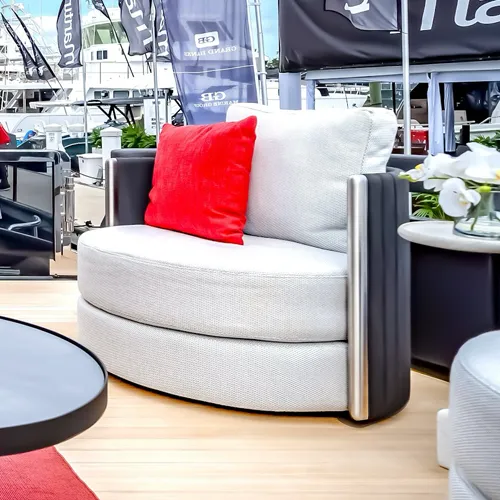 A light gray round outdoor armchair with dark armrests and a red pillow sits on a wooden deck. In the background, white yachts are docked under a bright sky.