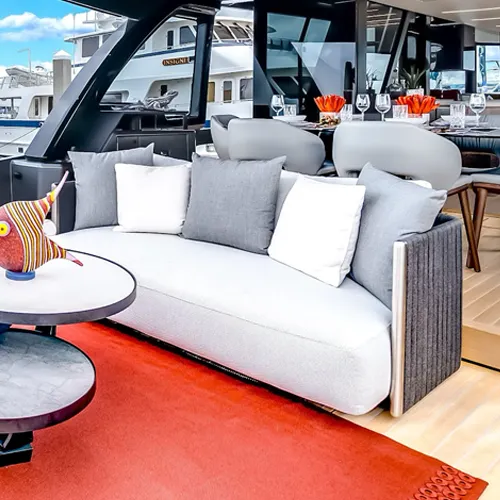 A luxurious outdoor seating area on a yacht with a light sofa, white and grey pillows, and two round tables. A red rug covers the floor, and other yachts are visible in the background.