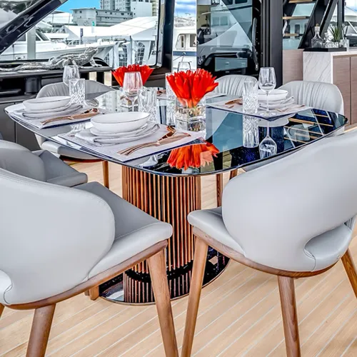 A modern dining table set for four with white plates, gold cutlery, and red flowers, overlooking a marina with boats through large windows.