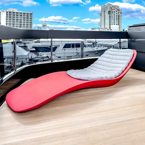 A red chaise lounge with a gray cushion on a wooden deck overlooking a marina with yachts and city buildings under a blue sky.
