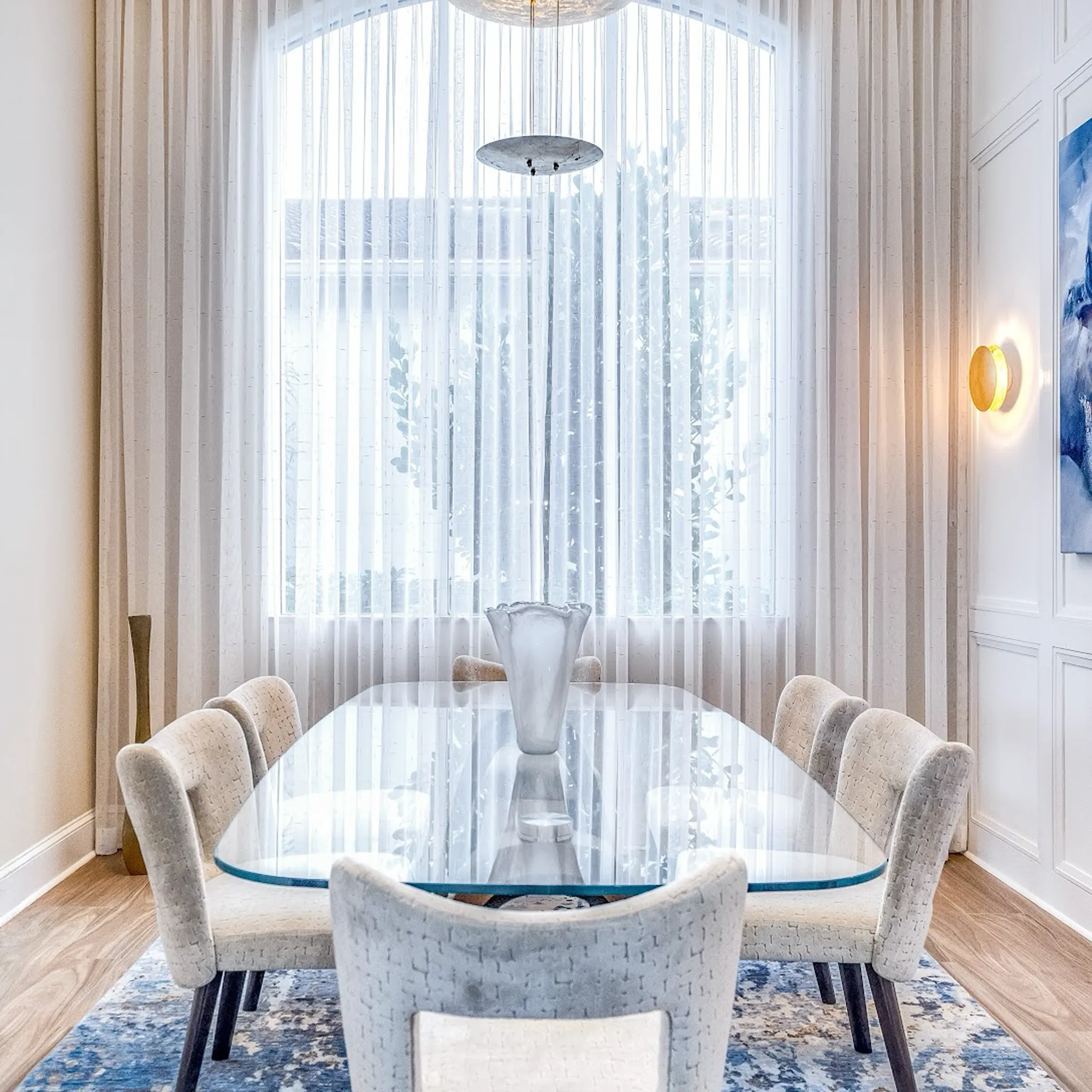 A modern dining room with an oval glass table, eight upholstered chairs, and a large arched window with sheer curtains. A blue and white rug covers the floor.