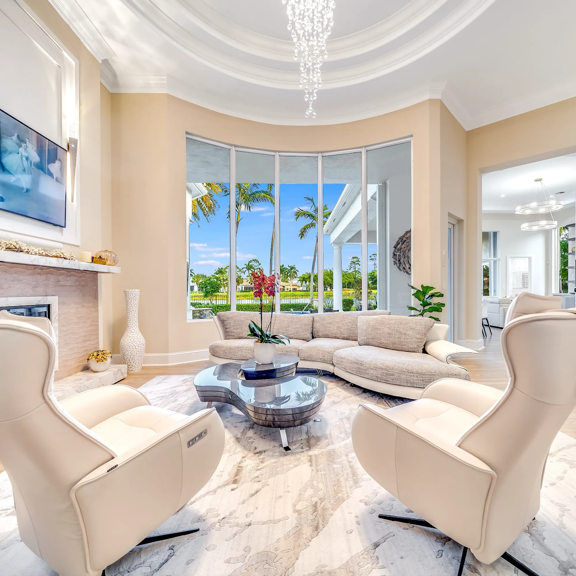 A modern, luxurious living room with a curved sectional sofa, two armchairs, a unique coffee table, and a fireplace, overlooking a tropical landscape through large bay windows.