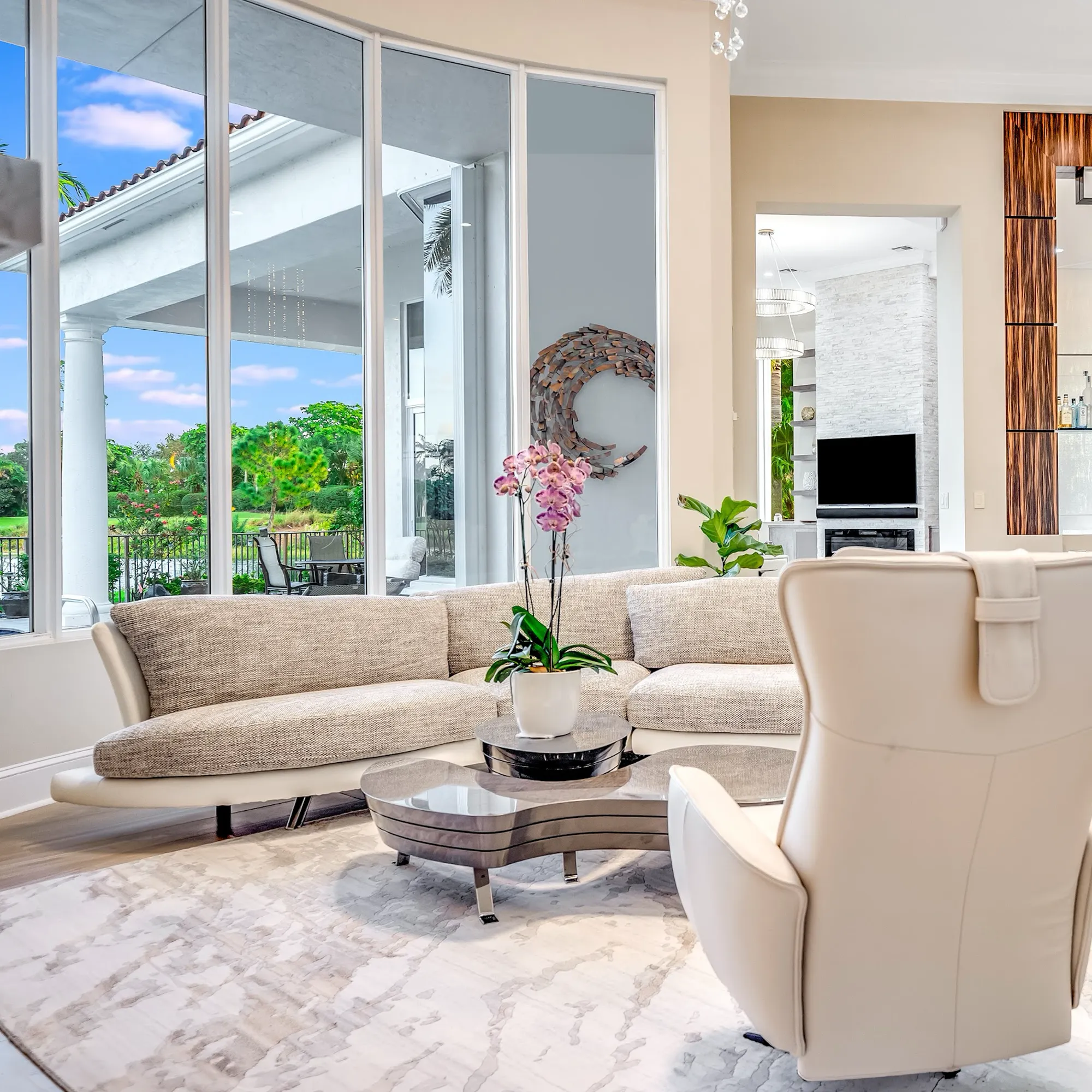 A modern living room with an L-shaped sofa, coffee table with an orchid, and large windows overlooking a lush green landscape and patio.
