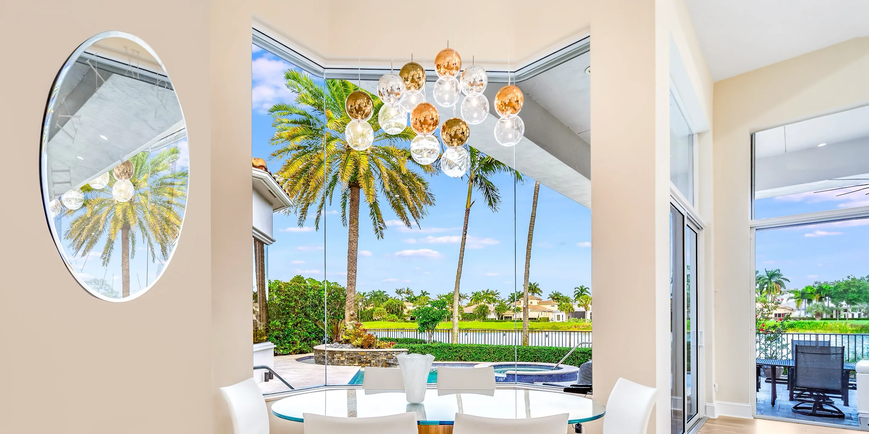 A dining room with a glass table and white chairs, featuring a modern spherical chandelier and large windows looking out to a tropical pool, palm trees, and a waterfront.