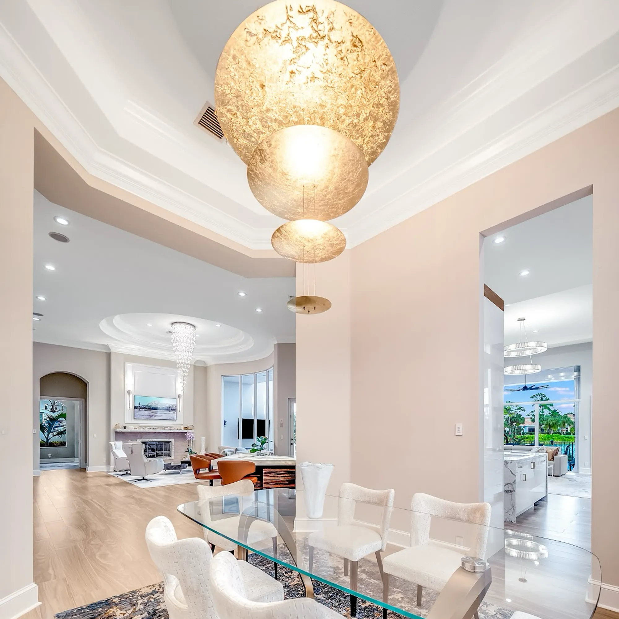 A luxurious open-concept home interior, showing a dining area with a glass table and white chairs, a living room with a fireplace, and a kitchen with an outdoor view. A multi-tiered gold chandelier hangs above the dining table.