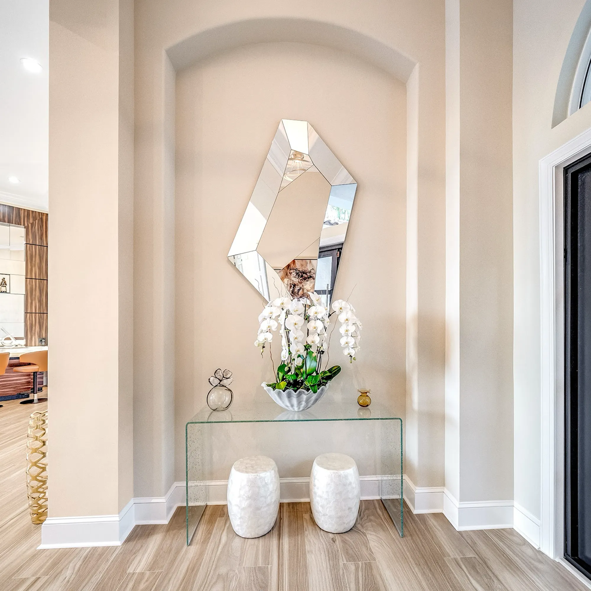 A modern entryway with a geometric mirror, glass console table, white orchids, and two white stools in an arched wall recess.