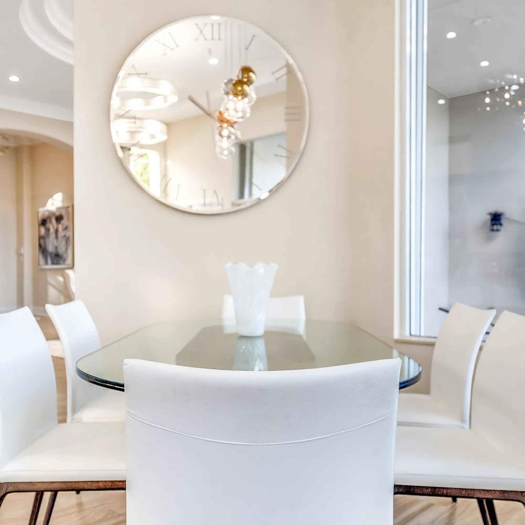 A modern dining room with a glass table, four white chairs, a white vase, and a large circular mirror clock on a beige wall.