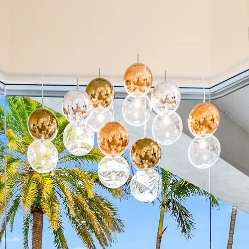 A modern chandelier with gold, rose gold, and clear spherical lights hangs in front of a window overlooking palm trees and a blue sky.