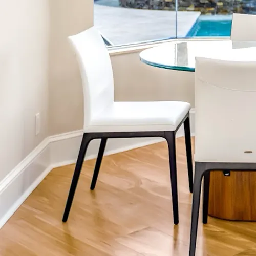 A white dining chair with dark legs next to a round glass table on a wood floor, with a window showing a pool.