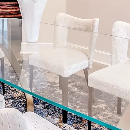 A clear glass dining table with a green edge, surrounded by light-colored upholstered chairs on a blue and white patterned rug.