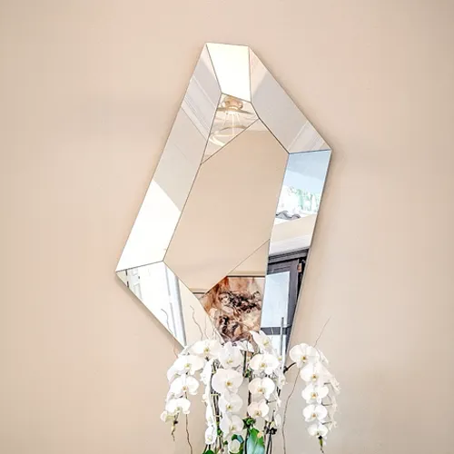 A modern, geometric mirror with an irregular hexagonal shape hangs on a light beige wall, reflecting parts of a room. Below it, a large arrangement of white orchids is visible.